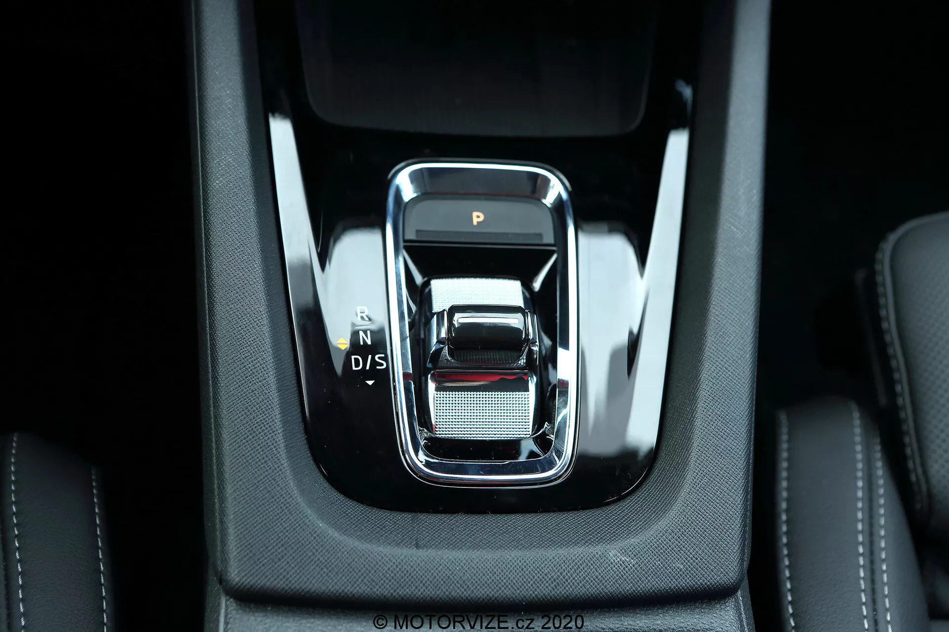 Detailný pohľad na modernú radiacu páku v stredovej konzole Škoda Octavia Limousine 2020. Predný pohľad na páku s elegantným, obdĺžnikovým dizajnom, zaoblenými hranami a chrómovým obrysom, zobrazujúcim polohy prevodov 'P', 'N', 'D/S'. Obklopený vysoko kvalitnými čiernymi materiálmi s viditeľným šitím a strieborným povrchom s jemnou sieťovou textúrou, naznačujúcimi luxus a detaily. Jasné osvetlenie osvetľuje robustný, konzistentný interiér, poukazujúci na pokročilé elektronické funkcie a moderné automobilové technológie.