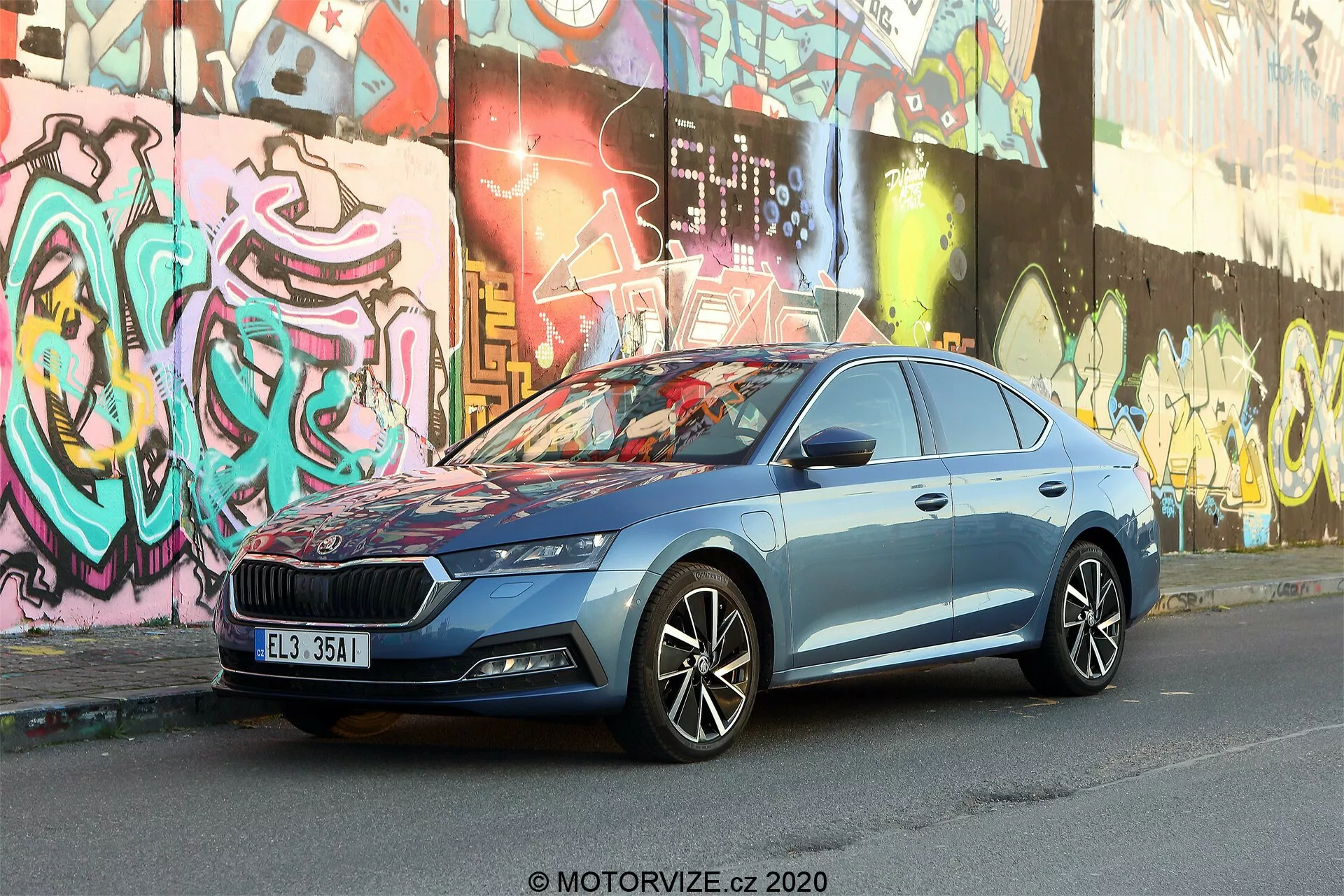 Obrázok zobrazuje Škoda Octavia Limousine 2020 zaparkovanú na mestskej ulici so stenami pokrytými graffiti v pozadí. Zachytené z mierne nízkeho predného trojštvrťového uhla, zdôrazňuje masívnu mriežku s vertikálnym vzorom a centrálne umiestneným emblémom, elegantné LED svetlomety, viaclúčové zliatinové kolesá, dynamické línie karosérie, bočné zrkadlá s integrovanými smerovkami, chrómované okenné lišty a kovovo modrú farbu. Mestské prostredie zdôrazňuje moderný dizajn a pokročilú automobilovú technológiu vozidla.