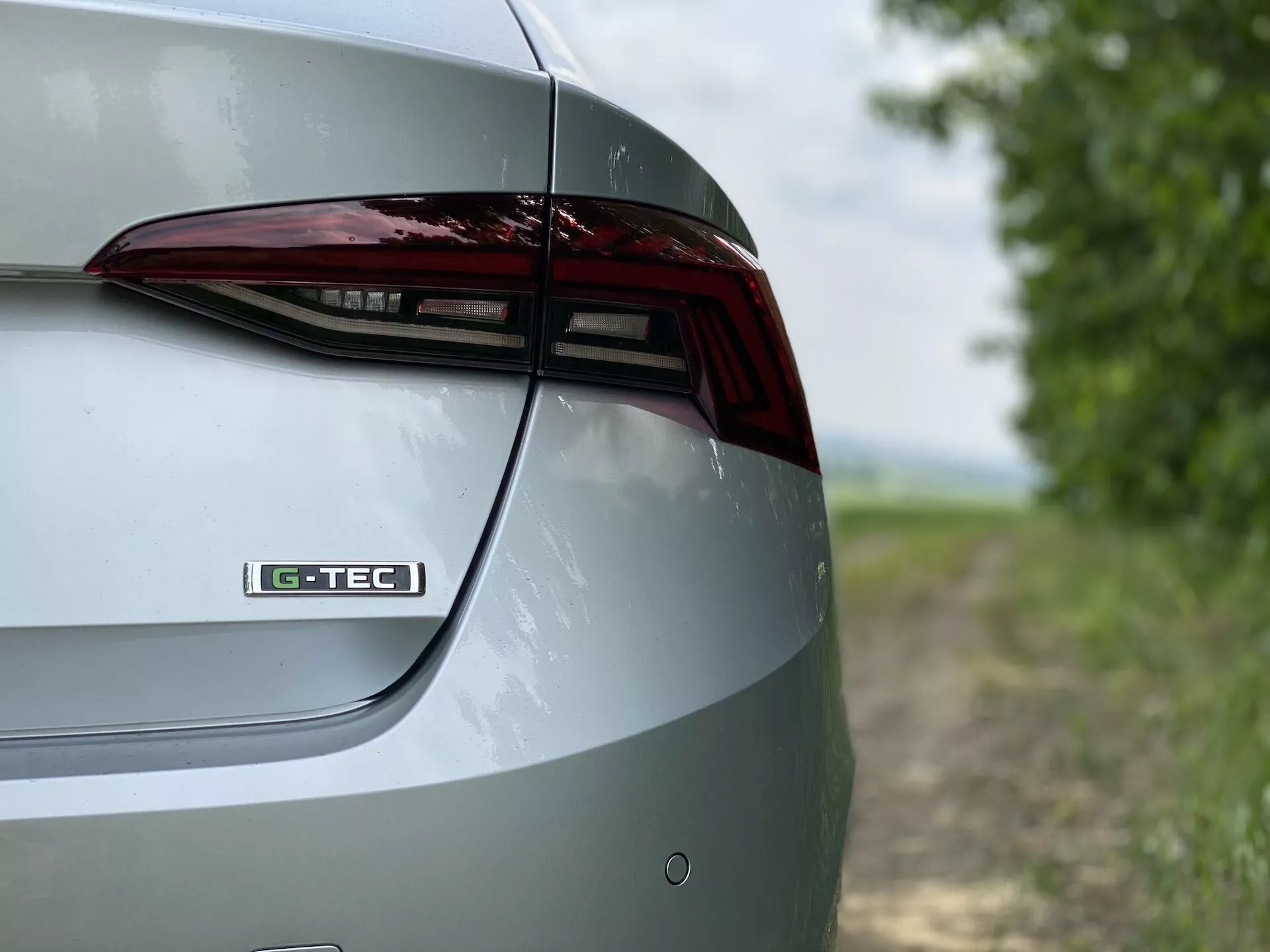 Detailný záber na pravú zadnú časť vozidla Škoda Octavia 2020 limuzína. Záber zachytáva dizajnové prvky veku kufra, LED zadného svetla a časti zadného nárazníka z šikmého, mierne vyvýšeného uhla. Odznak 'G-TEC' na veku kufra naznačuje špecifickú technológiu alebo výbavu. Kovový náter zvýrazňuje leštený exteriér vozidla. Rozmazané pozadie vytvára vidiecke prostredie, ktoré zvýrazňuje vlastnosti a dizajn vozidla.