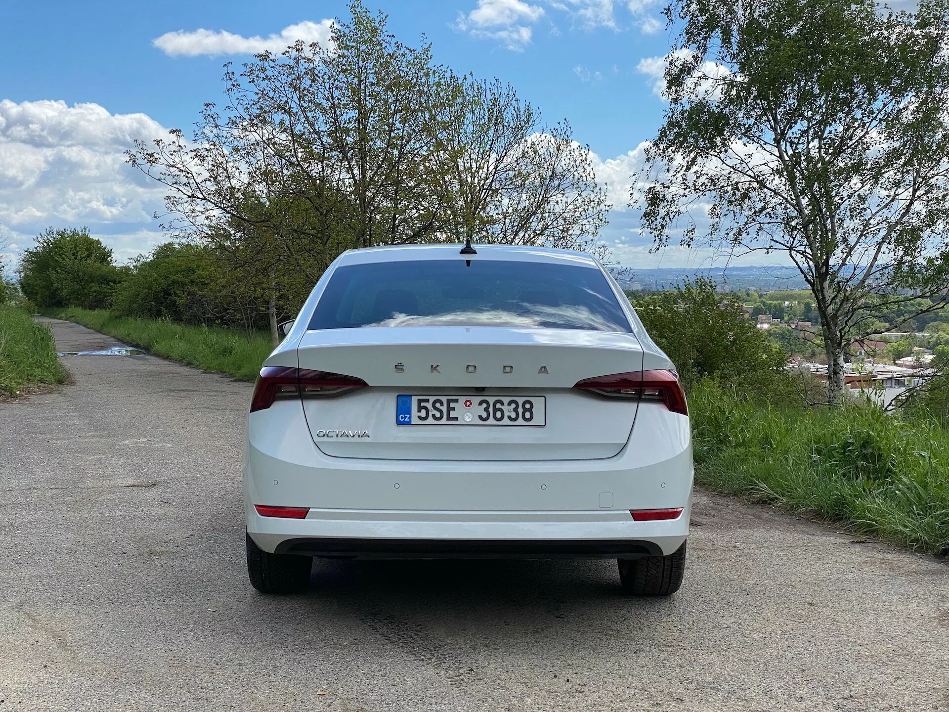 Pohľad na zadnú časť bieleho modelu Škoda Octavia 2020 limuzína, stojaceho na úzkej asfaltovej ceste v vidieckom prostredí. Obrázok, urobený z úrovne zeme priamo zepredu, ukazuje veko batožinového priestoru, zadný nárazník, zadné svetlá, poznávaciu značku, zadné sklo a logo Škoda umiestnené naprieč stredu veka batožinového priestoru s nápisom „Octavia“ na ľavo. Vozidlo obklopujú stromy a zamračená modrá obloha, s viditeľným vzdialeným mestom alebo krajinkou pod horizontom.
