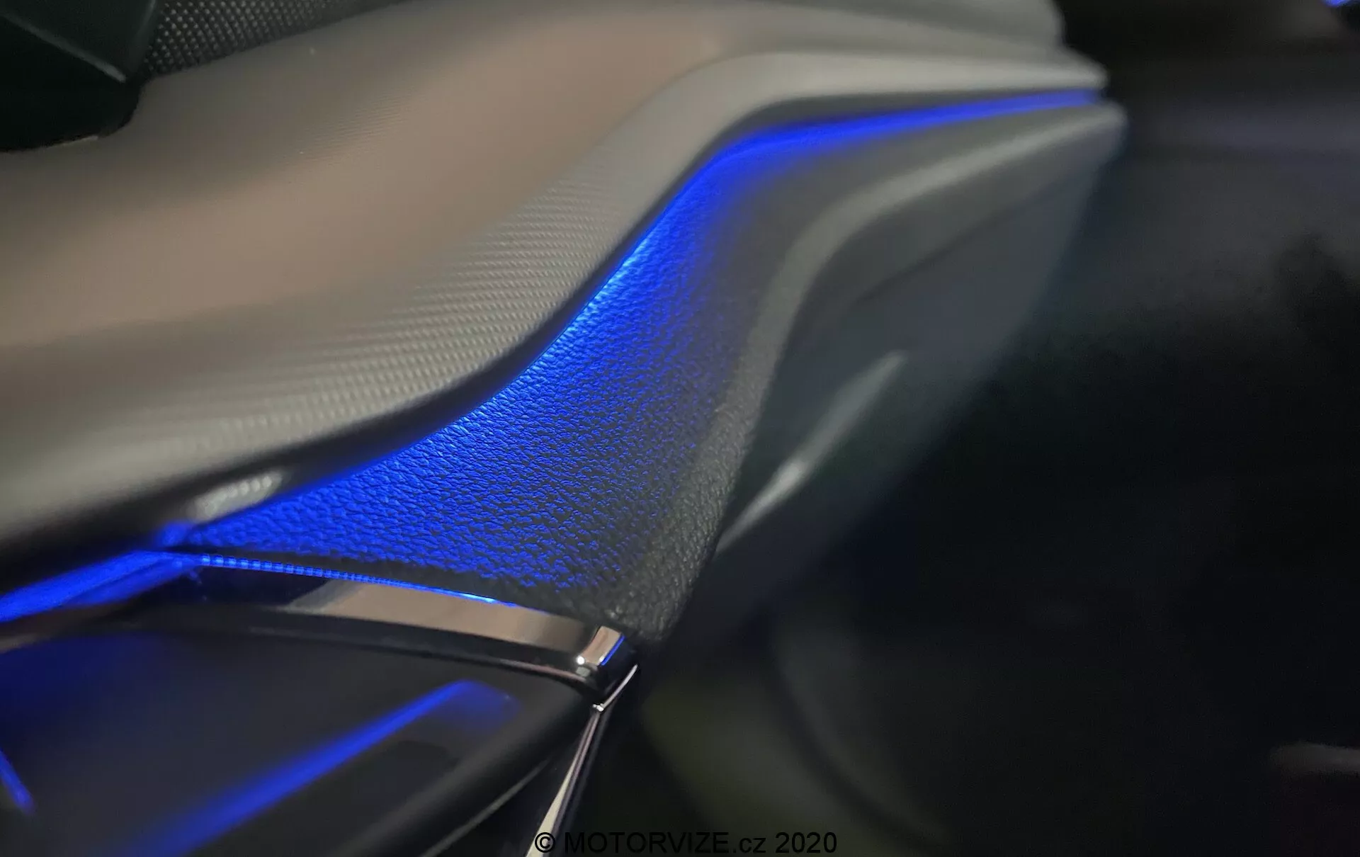 Detailný záber interiéru Škody Octavia Limousine 2020, zvýrazňujúci modrý prúžok ambientného osvetlenia, ktorý prebieha pozdĺž dverí alebo palubnej dosky vozidla. Elegantný dizajn s jemnými krivkami plynule splýva s povrchom kože, čím zdôrazňuje luxusný pocit pod jemným osvetlením. Fotografované v tmavom prostredí, snímka zvýrazňuje modré osvetlenie a textúry materiálov, predvádzajúce technológiu ambientného osvetlenia často ovládanú prostredníctvom infotainmentového systému vozidla.