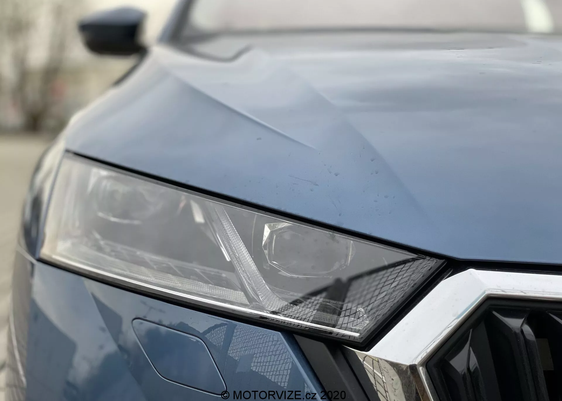 Detailný pohľad na prednú časť modelu Škoda Octavia 2020. Tmavá farba, zobrazujúca ľavý svetlomet s elegantným uhlovým dizajnom naznačujúcim technológiu LED alebo HID, geometrický šesťuholníkový vzor mriežky a uhlové záhyby na kapote pre športový vzhľad.