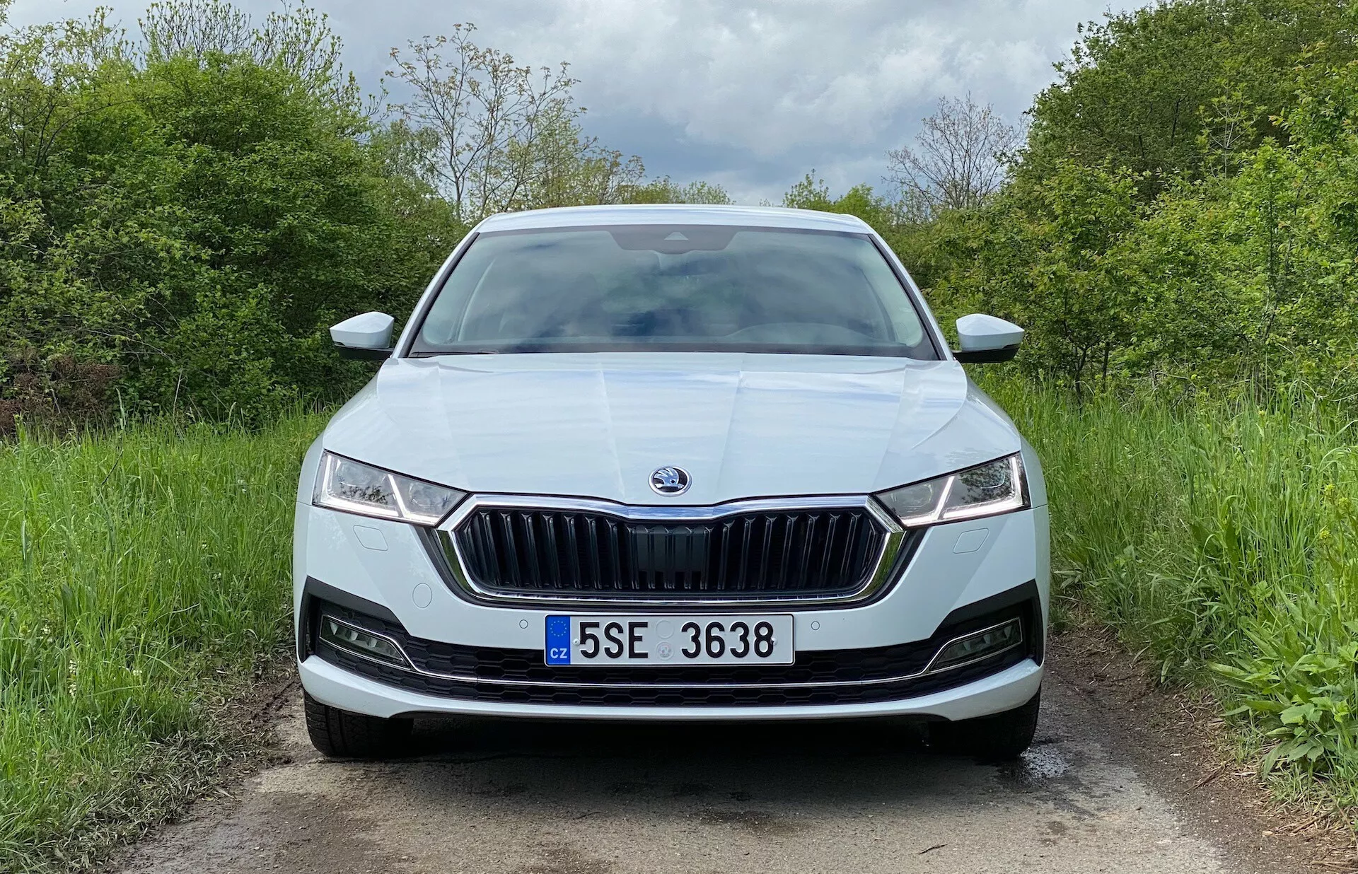 Predný pohľad na sedan Škoda Octavia 2020 na vidieckej ceste so zreteľnou prednou maskou s vertikálnymi lamelami a chrómovým lemom, úzkymi LED svetlami s denným osvetlením, centrálnym znakom značky, parkovacími senzormi v nárazníku, centrálnou registračnou značkou, metalickým lakom, nasávacími otvormi a možnými hmlovými svetlami.