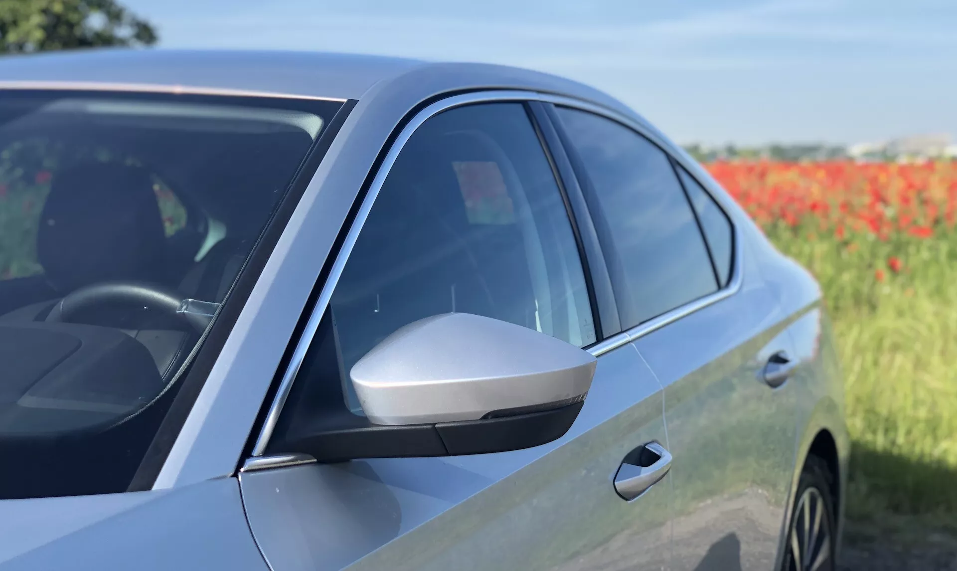 Detailný bočný pohľad na moderný sedan Škoda Octavia 2020, zameraný na predné dvere vodiča a bočné zrkadlo. Obrázok zdôrazňuje aerodynamické bočné zrkadlo s integrovaným smerovým svetlom, tónované okná, lesklý lak a aerodynamický dizajn od stredu k prednej časti. Odrážanie na paneli dverí a časť predného kolesa naznačujú možné zliatinové kolesá. Fotografia zdôrazňuje pokročilé dizajnové prvky a potenciálne technológie vozidla na mierne rozmazanom prírodnom pozadí.
