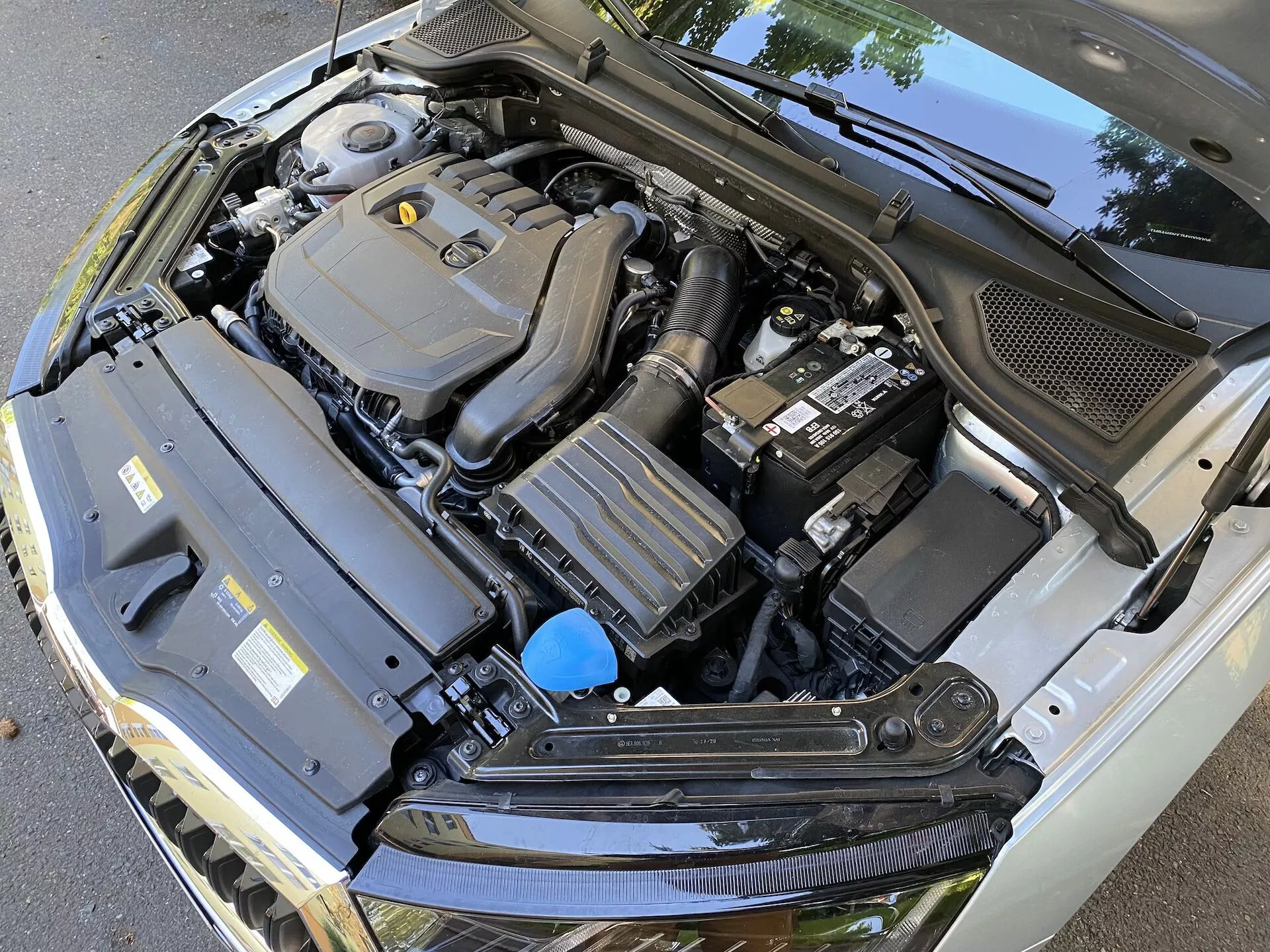 Detailný pohľad na motorový priestor vozidla Skoda Octavia 2020 limuzína s otvorenou kapotou, zobrazujúci rôzne komponenty, vrátane centrálne orámovaného krytu motora, batérie na pravej strane, obdĺžnikového komponentu s chladiacimi rebrami vľavo a nádrže na tekutiny s modrým uzáverom. Obrázok, nasnímaný z klesajúceho, mierne vycentrovaného uhla, tiež zobrazuje čierny plastový kryt na spodnej hrane čelného skla a zvýrazňuje technickú zložitosť motorového priestoru automobilu.