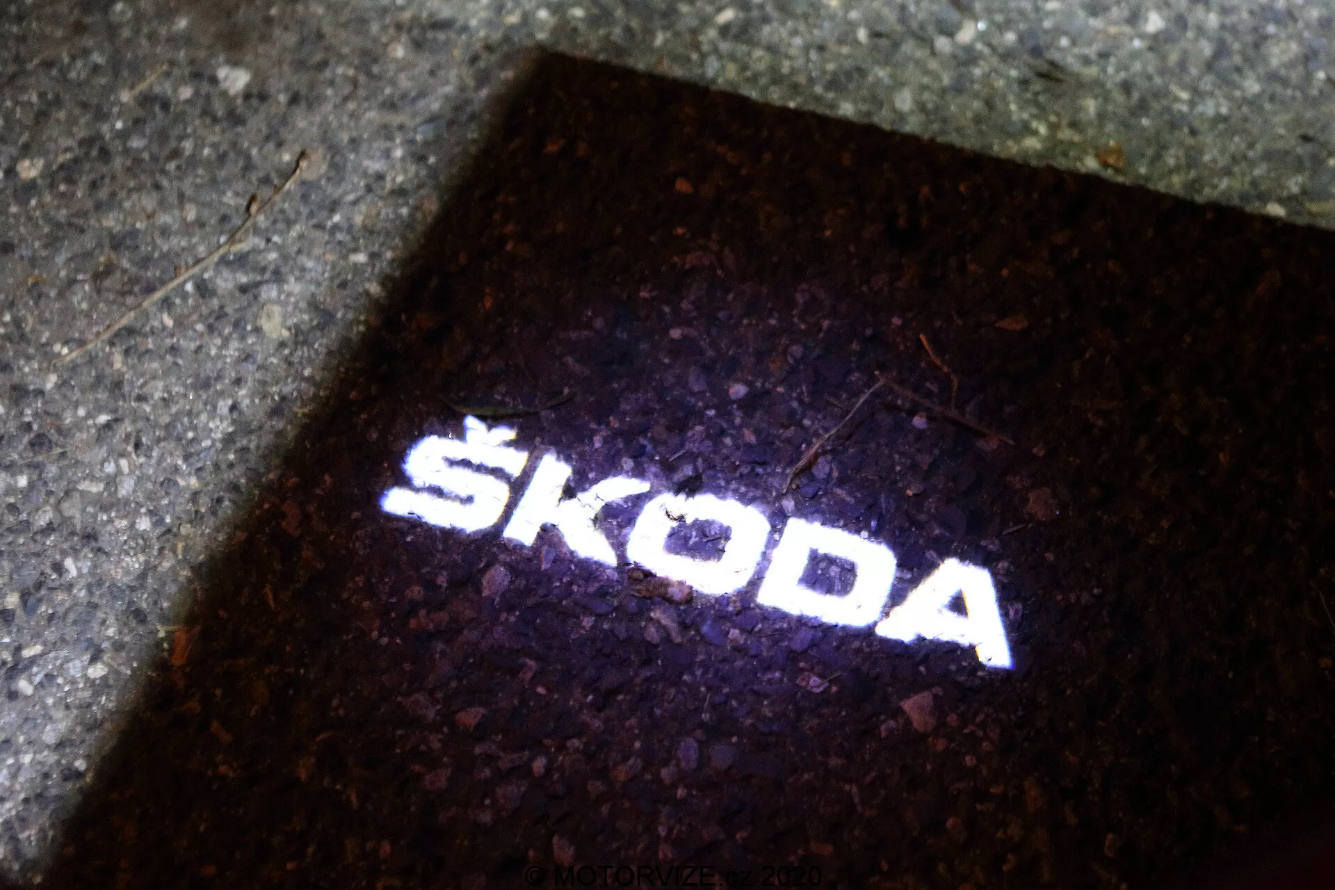 Projekcia loga Škoda na zem. Zdroj svetla, pravdepodobne umiestnený na exteriéri vozidla Škoda Octavia 2020 (octavia-limousine), vytvára 'puddle light', ktorý osvetľuje zem pri otvorení dverí vozidla. Táto ostrá a jasná biela projekcia svetla na tmavom pozadí slúži na funkčné aj brandingové účely a je indikáciou prítomnosti modernej LED technológie v príslušenstve vozidla.
