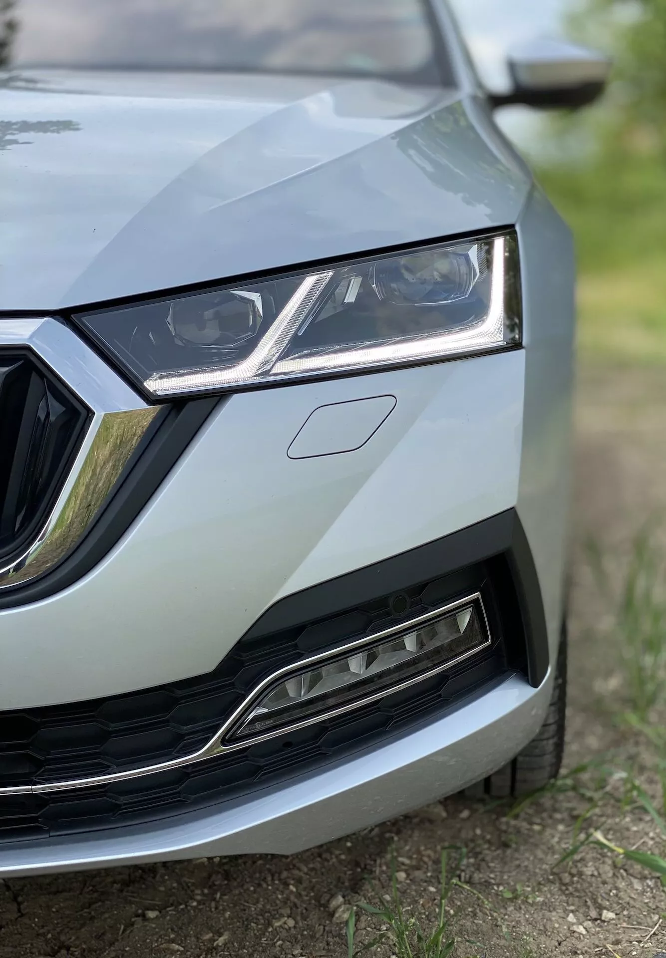 Detailný záber na prednú ľavú časť Škody Octavia 2020 (Octavia Limousine) zobrazujúci detaily od stredu mriežky po ľavý svetlomet, časť kapoty a časť predného nárazníka a blatníka. Snímka urobená z nízkeho uhla zvýrazňuje elegantný dizajn svetlometu s LED denným osvetlením, chrómovo zdobenú mriežku a dolnú mriežku s moderným, kovovým povrchom. Zaostrenie je na detaily vozidla s nízkou hĺbkou ostrosti, čo vytvára profesionálny, vysokokvalitný vzhľad.