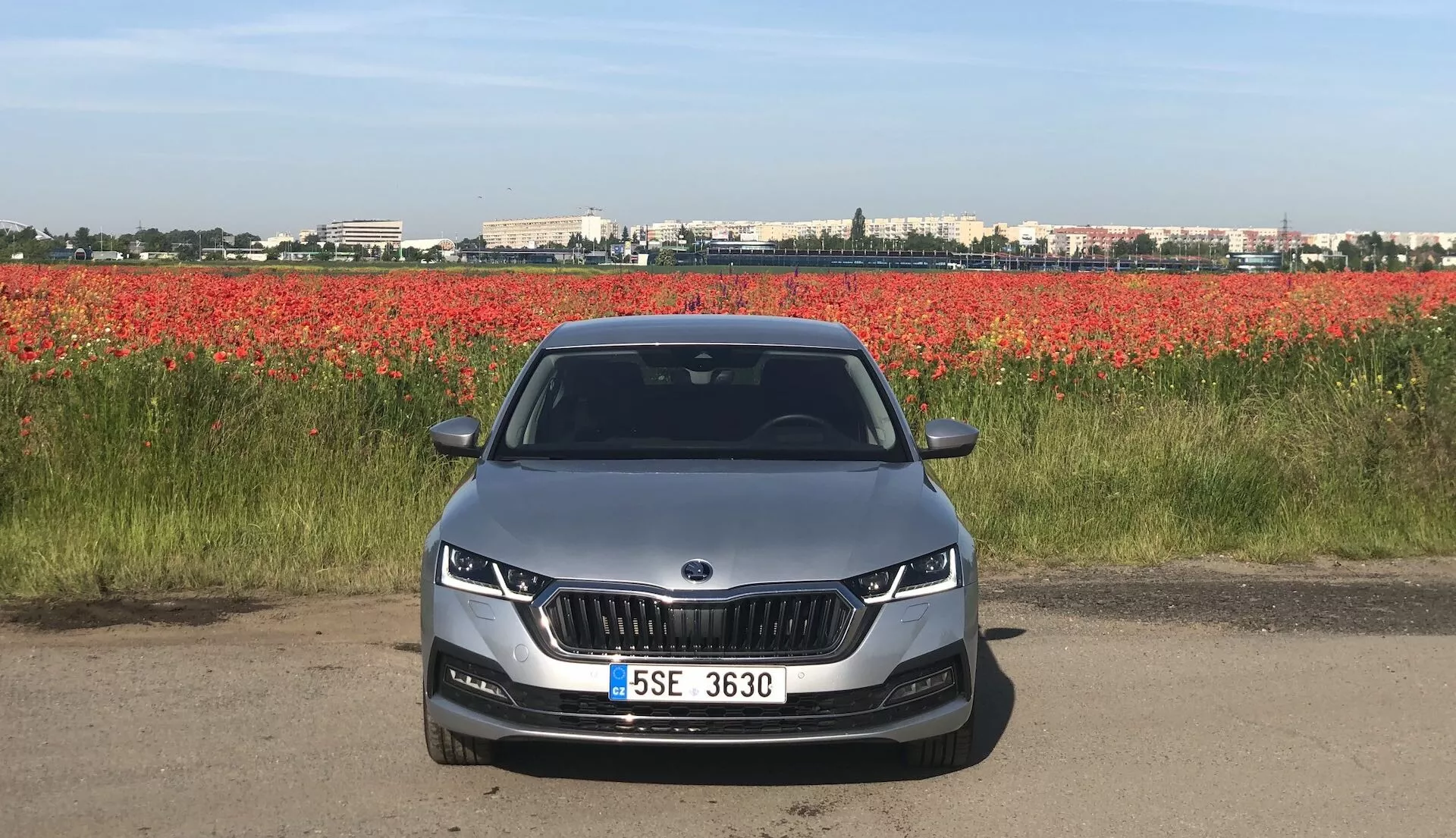 Predný pohľad na Škoda Octavia Limousine 2020 na ceste alebo štrkovej ceste, predvádza svoju prednú časť s charakteristickou mriežkou v tvare obličky, obklopenou elegantnými LED alebo adaptívnymi svetlami. Aerodynamický predný nárazník má výrezy pre hmlové svetlá a vzduchové prieduchy. Kapota má kontúrové línie pre estetický a aerodynamický dizajn. Plne viditeľné čelné sklo môže naznačovať pokročilé technologické prvky ako head-up displeje alebo asistenčné systémy vodiča. Svetlý automobil s centrálne umiestnenou ŠPZ a parkovacími senzormi, na pozadí červených kvetov.