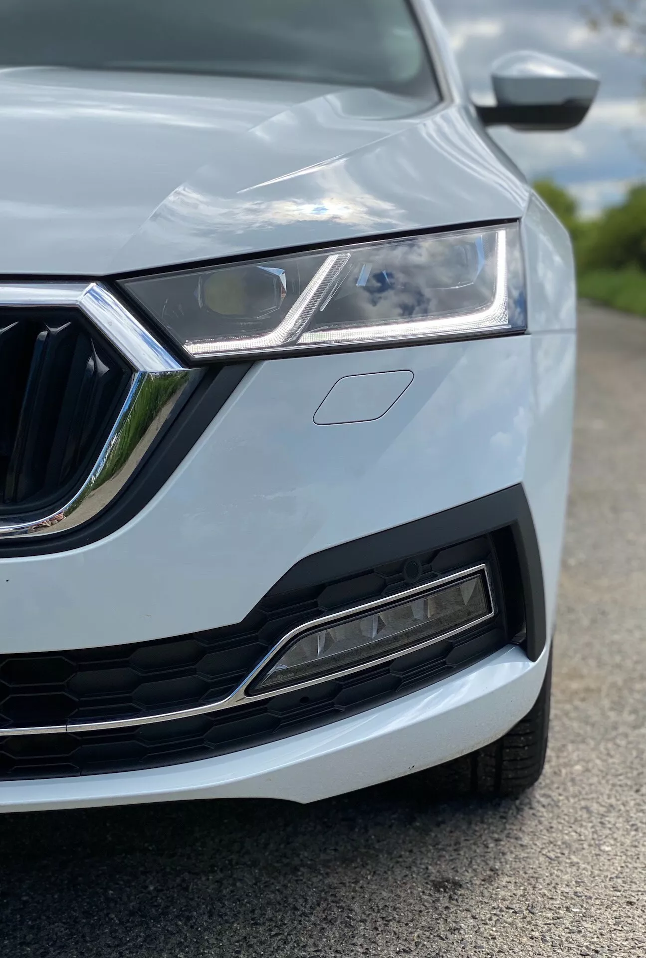 Blízky pohľad na ľavú prednú štvrtinu vozidla Škoda Octavia 2020 (octavia-limousine), ktorý zvýrazňuje LED svetlomet, charakteristickú mriežku s chrómovými detailmi, moderný predný nárazník s prívodom vzduchu a senzormi, dizajn kapoty s ohybom a čiastočne viditeľné spätné zrkadlo s integrovanými smerovými svetlami. Auto je natreté svetlou farbou, pravdepodobne bielou alebo striebornou, so štíhlymi líniami karosérie. Obraz je nasnímaný z nízkeho uhla, zameriava sa na svetlomet a mriežku, s plytkou hĺbkou ostrosti, ktorá rozmazáva okolité prvky.