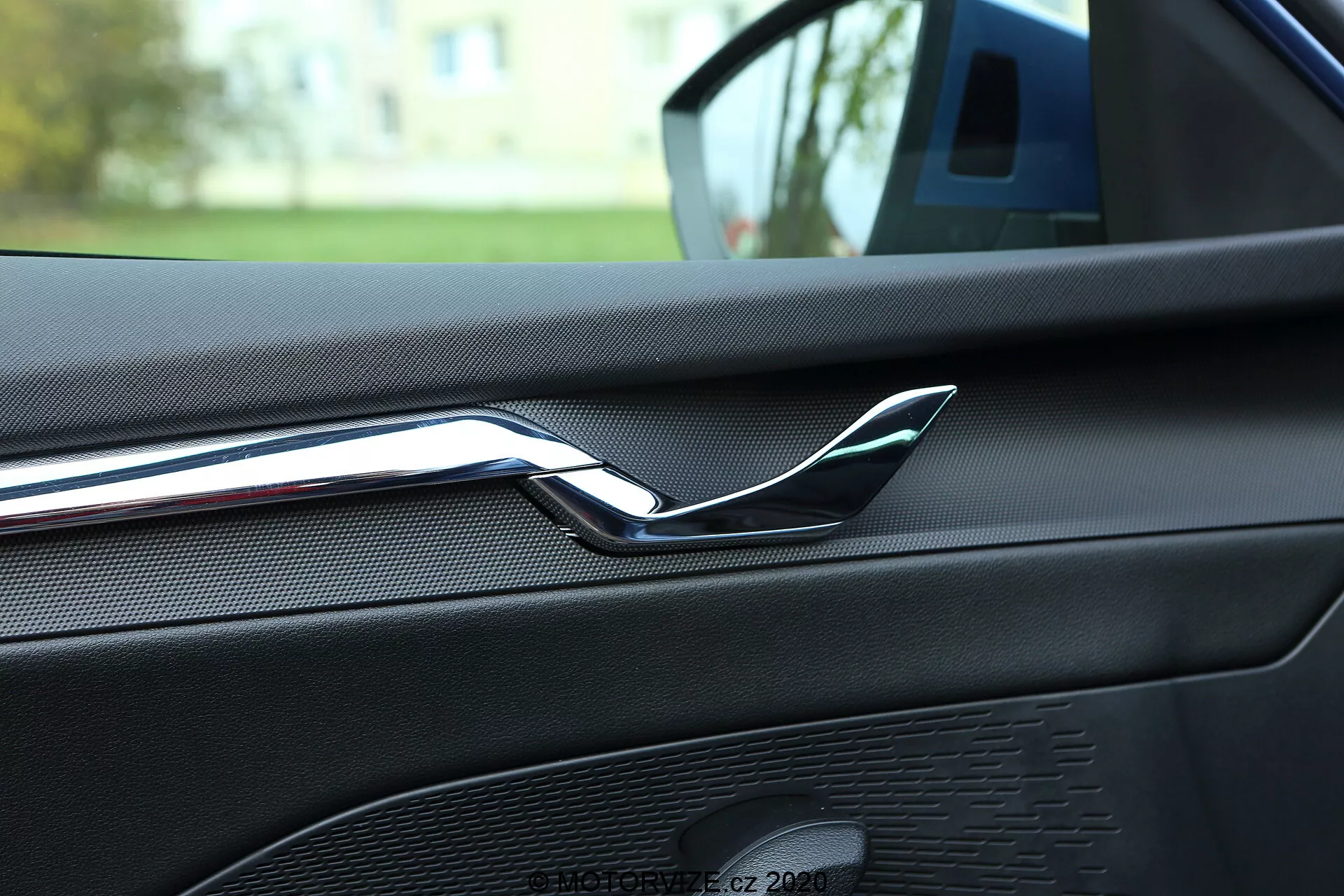 Detailný pohľad na interiér Škoda Octavia 2020, zameraný na zadnú časť predných dverí spolujazdca, zvýrazňujúci reflexnú chrómovanú kľučku dverí. Dverový panel je potiahnutý koženkovým alebo štruktúrovaným mäkkým materiálom s viditeľným šitím, čo naznačuje vysokokvalitnú povrchovú úpravu interiéru. Tiež viditeľná mriežka reproduktora ako súčasť audiosystému auta s jednotným vzorom a farebne zladeným dizajnom. Obrázok je urobený zo zadného sedadla, čo naznačuje, že auto je zaparkované vonku v obytnej oblasti, s jemne rozmazaným zeleným pozadím.