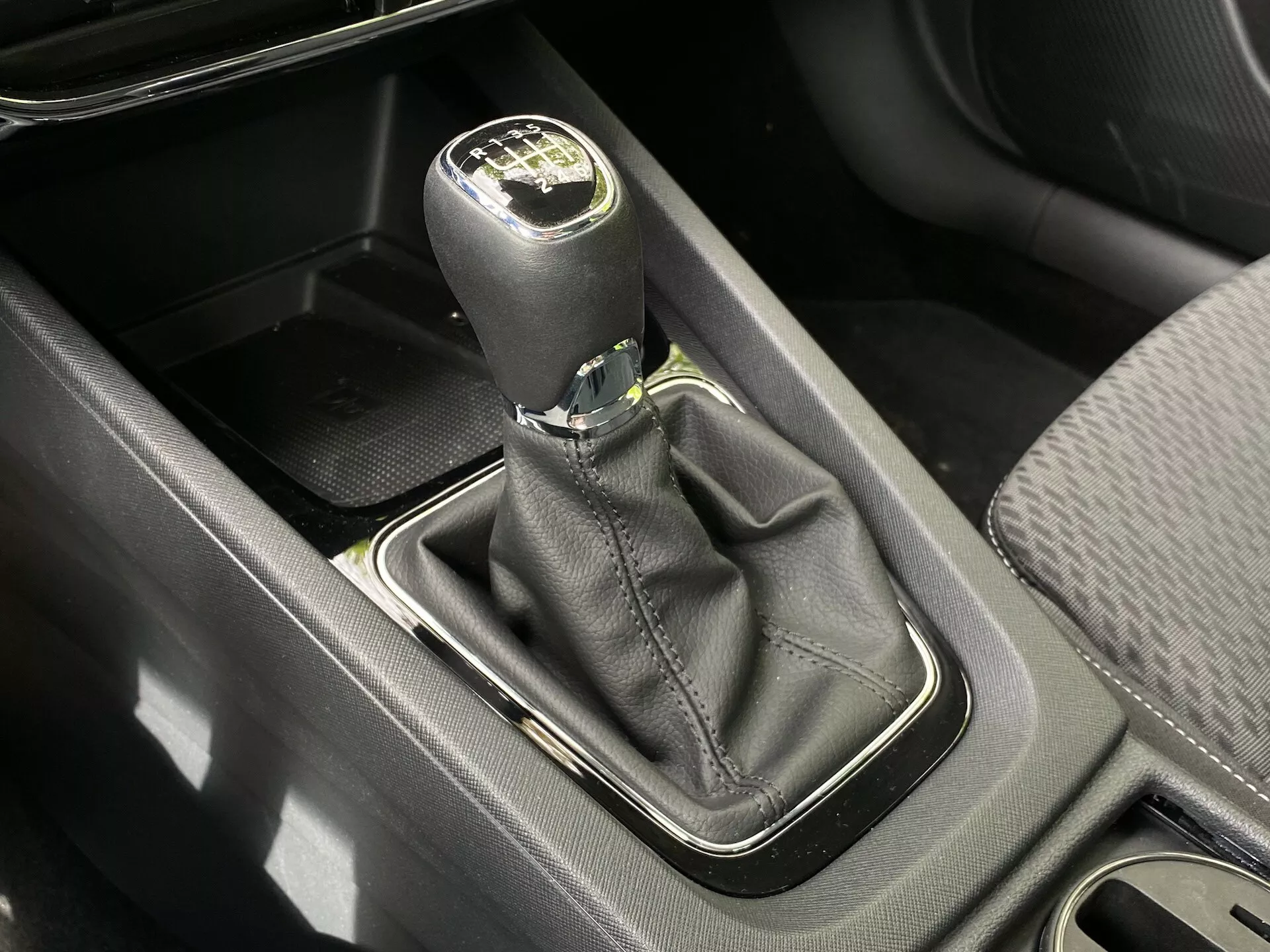 Pohľad z vtáčej perspektívy na radiacu páku s manuálnou prevodovkou v automobile Skoda Octavia 2020 (Octavia-Limousine). Čierna kožená radiaca páka so strieborným akcentom zobrazuje rozloženie prevodov pre šesťstupňovú prevodovku plus spiatočku. Základňa radiacej páky, obklopená strieborným rámčekom, je umiestnená v čiernej koženej manžete. Na obrázku sú tiež vidieť úložné priehradky, stredová konzola a držiak na nápoje, osvetlené denným svetlom.