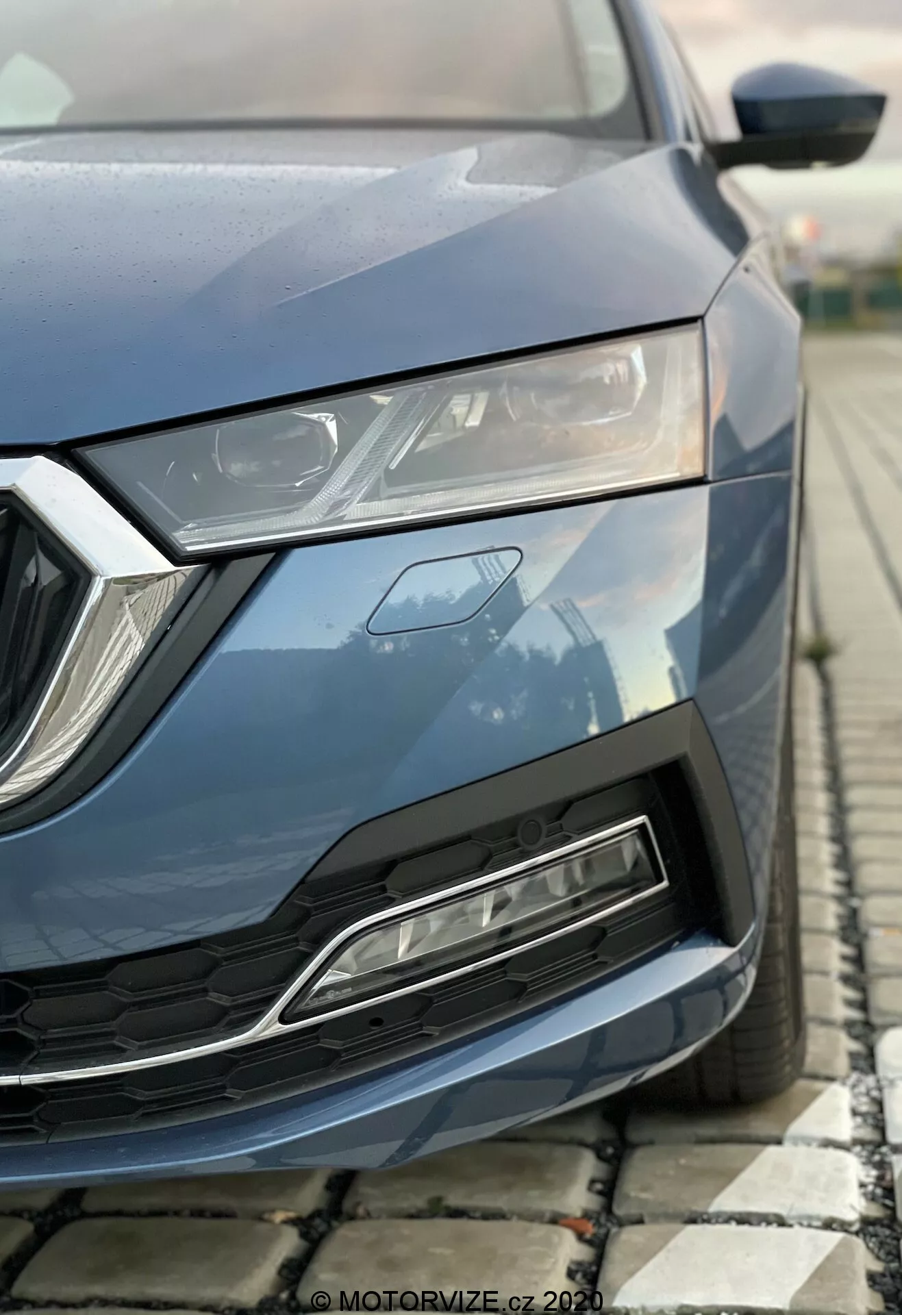 Blízky čiastočný obraz moderného auta Skoda Octavia 2020 zachytený z nízkeho uhla, zameraný na prednú-ľavú časť zahŕňajúcu kapotu, elegantný LED svetlomet, geometrickú mriežku a predný nárazník s prívodom vzduchu. Metalická modrá farba, uhlové dizajnové prvky a zdôraznené aerodynamické vlastnosti.