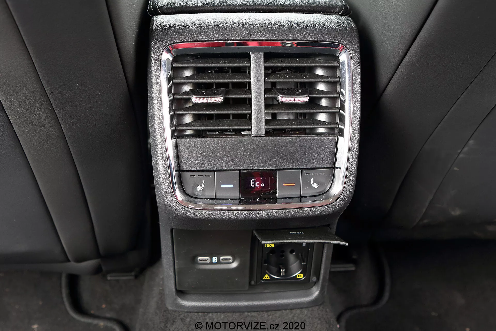 Obrázok zachytáva stredovú konzolu interiéru vozidla Škoda Octavia Limousine 2020, zdôrazňujúc ovládacie prvky klimatizácie a pohodlie zadných cestujúcich. Fotografia zreteľne ukazuje nastaviteľné vetracie otvory, tlačidlá na nastavenie teploty, tlačidlo režimu Eco, 12V zásuvku a úložný priečinok.