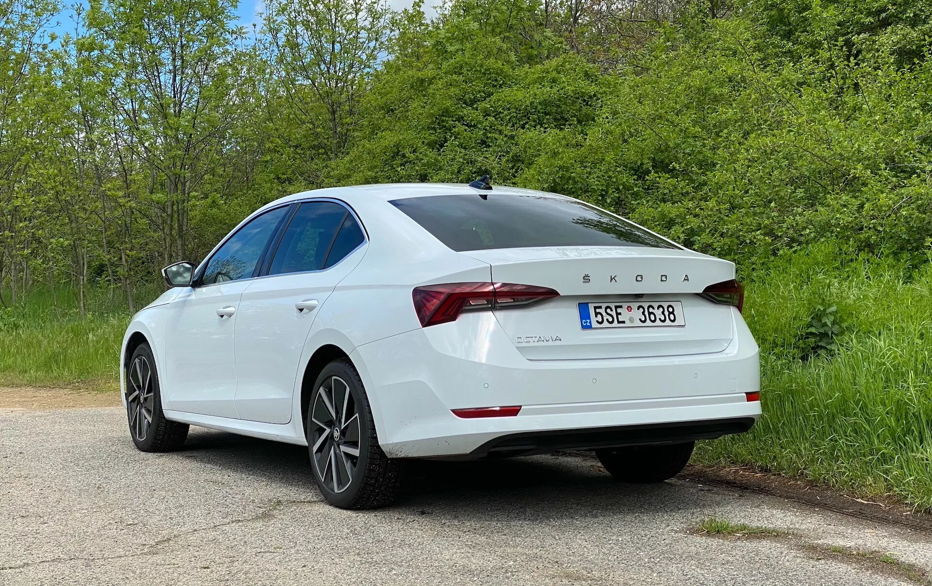 Obrázok zobrazuje biely sedan Škoda Octavia 2020 z uhla tri štvrte zadnej strany, zvýrazňujúci pravú stranu a zadné prvky vozidla. Kľúčové detaily zahŕňajú LED zadné svetlá, logo Škoda a označenie modelu OCTAVIA, nápis TDI, tvarovaný kryt batožinového priestoru so zabudovaným spojlerom, päťramenné zliatinové kolesá, hladkú, klesajúcu líniu strechy, zadný nárazník so senzormi parkovania a lesklý, dobre udržiavaný lak. Auto je zaparkované na pokojnej vidieckej alebo parkovej ceste so zeleňou na oboch stranách, čo umocňuje pokojné prostredie. Táto fotografia zdôrazňuje vonkajší dizajn auta, moderné prvky sedana a technologické prvky ako je dieselový motor a systémy asistencie vodiča.