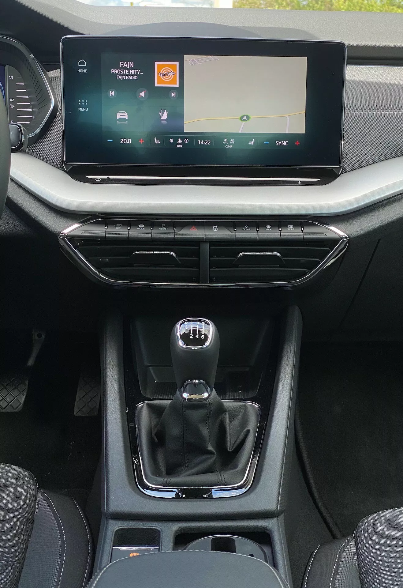 Detailný pohľad na stredovú konzolu interiéru Škoda Octavia limuzína 2020 z perspektívy zadného sedadla, zobrazujúci široký dotykový displej infotainmentu s ovládaním médií a klímy, vetracie otvory s kovovým akcentom a minimalistickými dizajnovými tlačidlami, automatickú radiacu páku v štýle tradičnej manuálnej radiacej páky so znamením manuálneho režimu, úložný priestor s USB portmi, držiaky na nápoje a zmes kožených a hladkých plastových štruktúr zdôrazňujúcich moderný komfort a zameranie na používateľskosť pre vodiča.