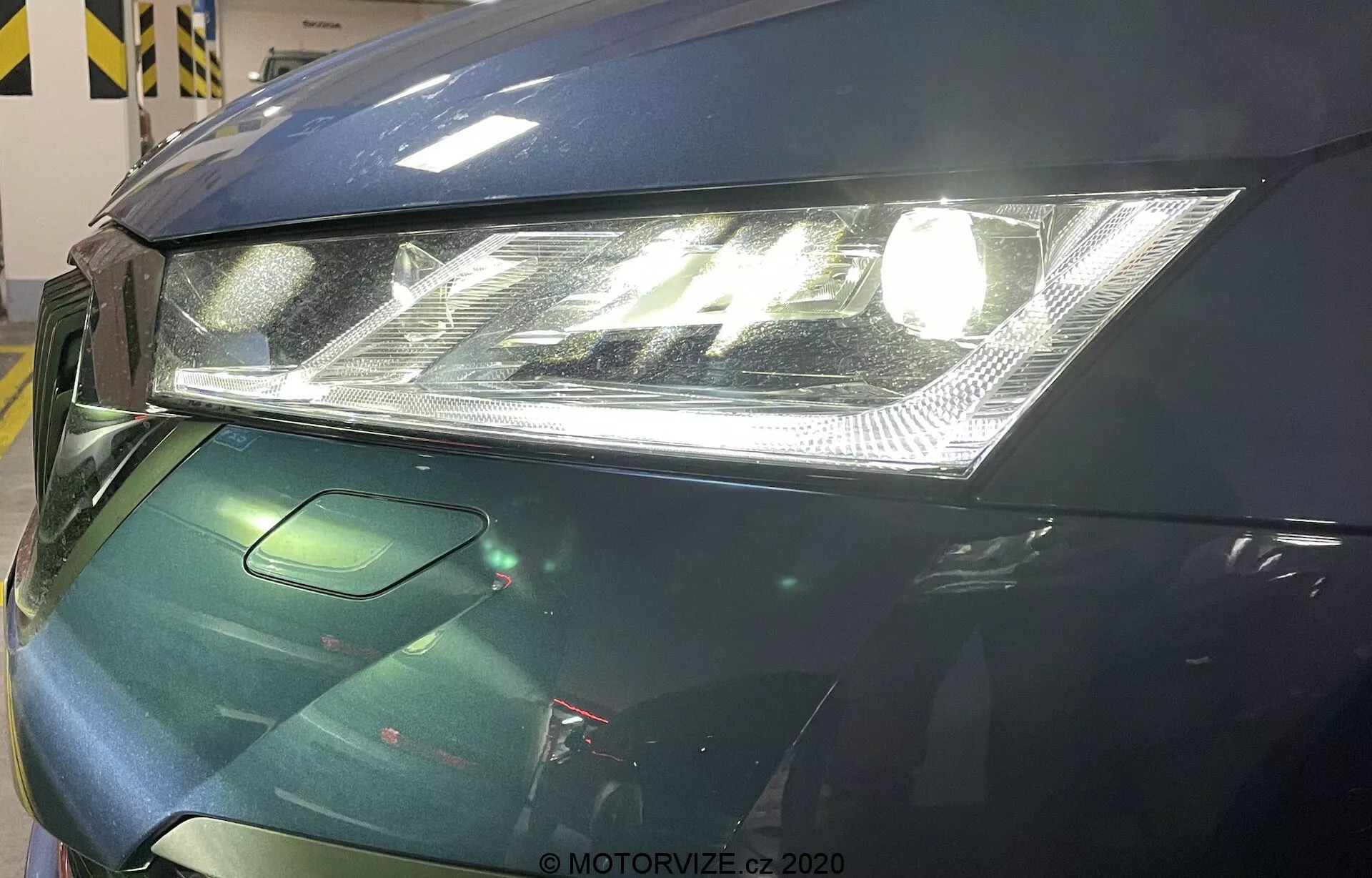 Detailný pohľad na prednú ľavú časť vozidla Škoda Octavia 2020. Obrázok sa zameriava na moderný svetlomet s čírou šošovkou a integrovanými LED dennými svetlami, čiastočne viditeľnú kapotu s hladkým dizajnom a malú časť mriežky, ktorá pravdepodobne obsahuje senzory vodičských asistenčných technológií. Vozidlo má tmavozelený lesklý povrch, čo naznačuje pokročilé opatrenia na ochranu laku.