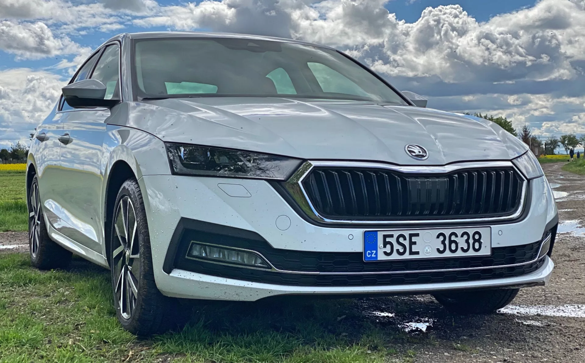 Pohľad z nízkeho uhla na prednú štvrtinu bielej Škodky Octavia 2020 zaparkovanej vonku, zdôrazňujúci prednú časť, ostrý dizajn svetlometov s LED denné osvetlenie, širokú mriežku s logom Škoda, parkovací senzor, športový predný nárazník s hmlovými svetlami, viaclúčové zliatinové kolesá a integrovaný smerovku v bočnom zrkadle. Viditeľné sú aj panoramatické čelné sklo a európska poznávacia značka.