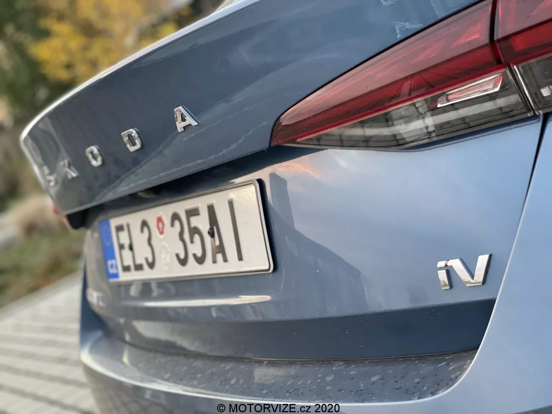 Detail zadnej časti modelu Škoda Octavia Limousine 2020, zachytený z nízkeho uhla so zameraním na veku batožinového priestoru, časť zadného nárazníka, vrátane chrómového loga Škoda, odznaku iV na hybridnú alebo elektrickú variantu, európskeho štýlu poznávacej značky, úzkych horizontálne orientovaných zadných svetiel zasahujúcich do veku batožinového priestoru a reflexného pruhu na zadnom nárazníku.