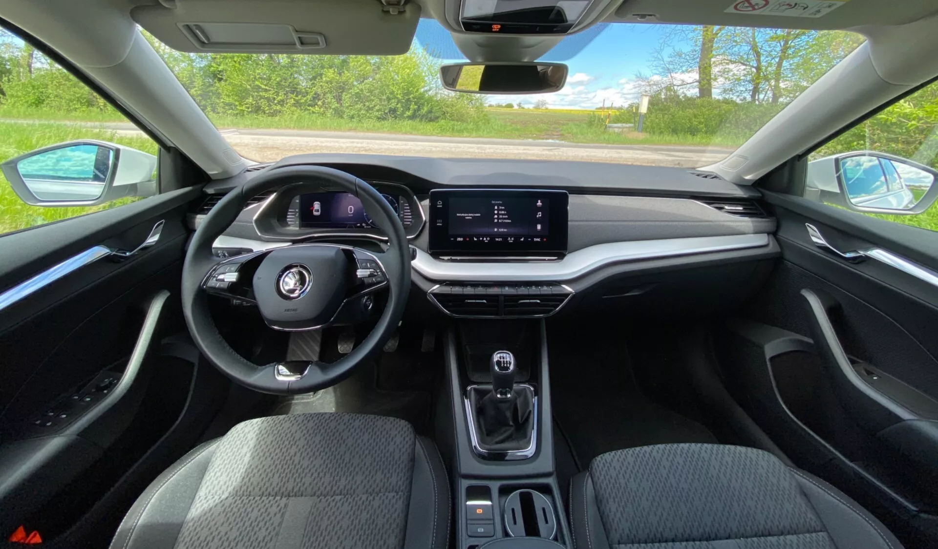 Interiér vozidla Škoda Octavia 2020 z pohľadu vodiča, zobrazujúci multifunkčný volant, digitálny prístrojový panel, horizontálne orientovanú infotainment dotykovú obrazovku, minimalistickú stredovú konzolu s voličom prevodovky, držiaky na nápoje a mäkkú palubnú dosku s dreveným dekorom za jasných denných podmienok.