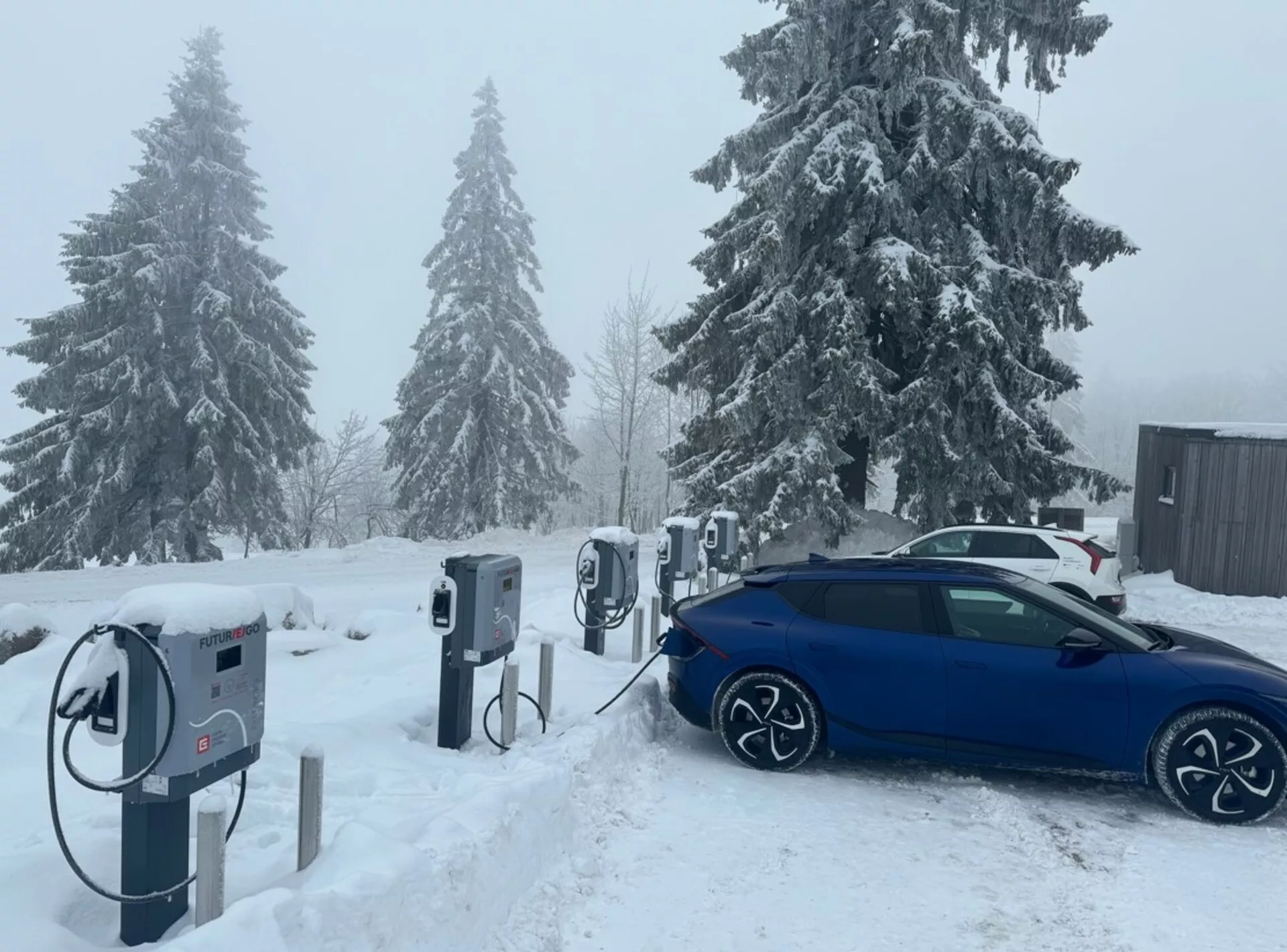 Modrá limuzína Škoda Octavia 2020 nabíjaná na stanici pre elektrické vozidlá, pohľad šikmo zo zadu, zdôrazňuje jej zliatinové kolesá, nabíjací port a dizajn pripravený na zimu v snehových podmienkach.