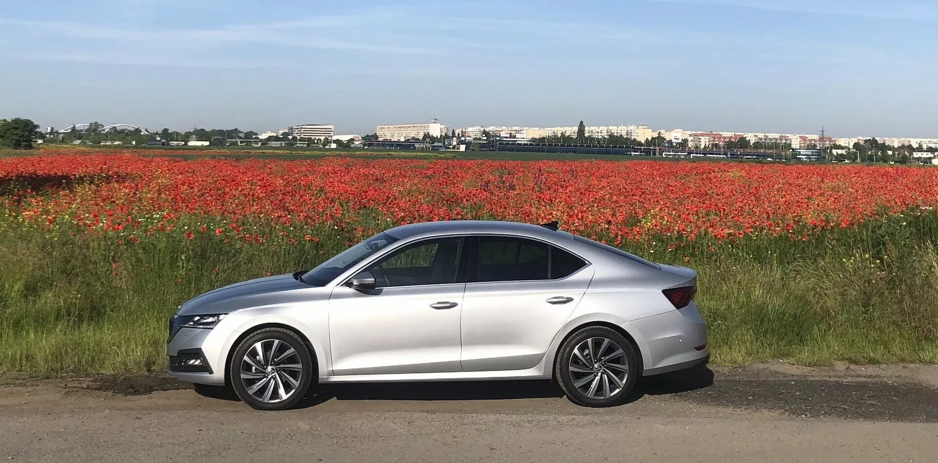 Bočný pohľad na moderný strieborný sedan Škoda Octavia 2020 zaparkovaný na rovnej ploche vedľa poľa červených kvetov, s elegantným aerodynamickým dizajnom, LED svetlometmi, bočnými zrkadlami farby karosérie s integrovanými smerovkami, zliatinovými kolesami, štyrmi dverami, tónovanými oknami a jemne sa zvažujúcou líniou strechy.