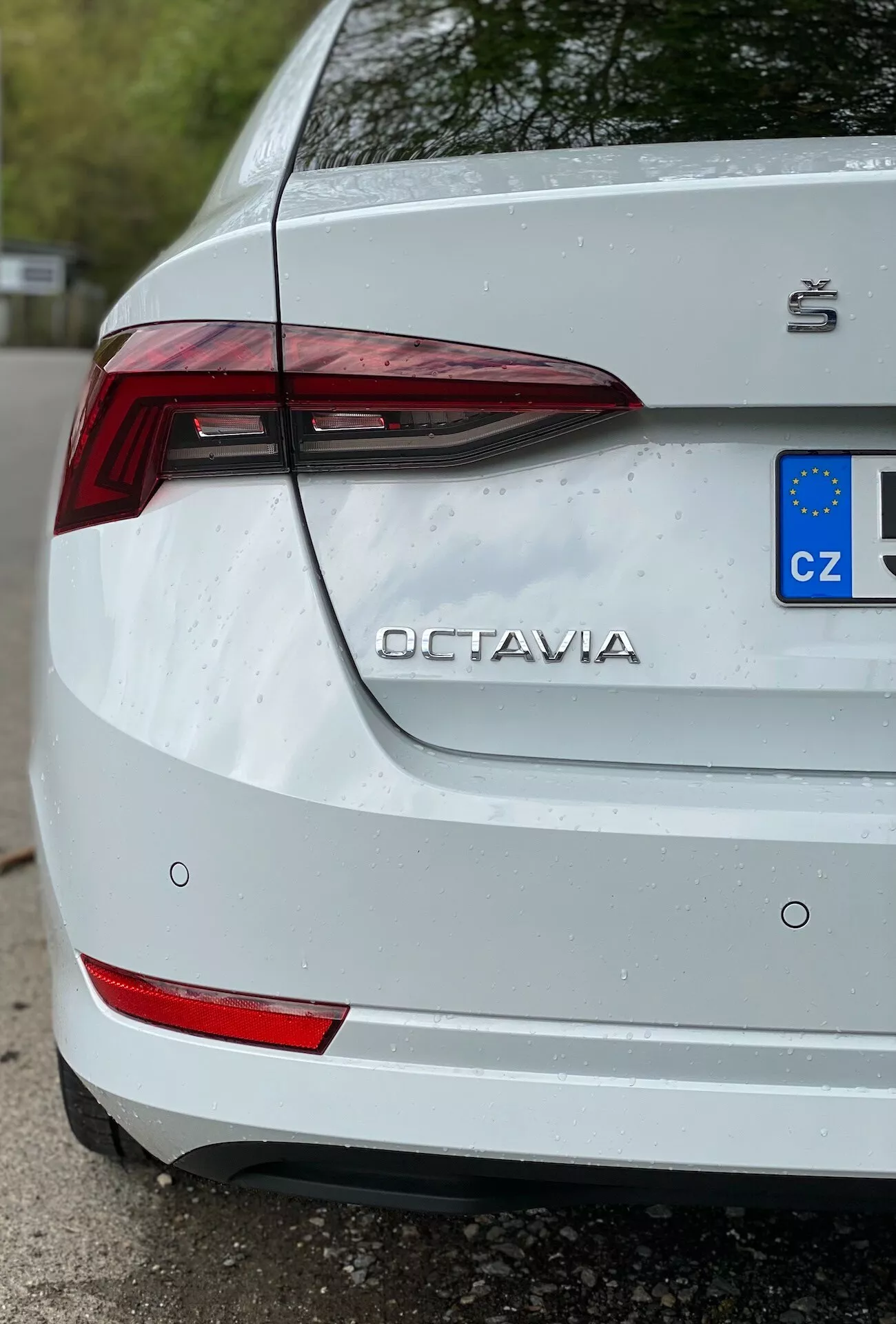 Detailný záber zadnej časti bielej Škody Octavia 2020 (octavia-limousine) z trojštvrťovej perspektívy zameranej na pravú stranu. Obrázok zdôrazňuje významné črty, ako sú ostré LED zadné svetlo, odrazové prvky na zadnom nárazníku, názov 'OCTAVIA' a logo Škoda na veku kufra a čiastočná európska evidenčná značka s označením 'CZ'. Jemné kvapky vody na povrchu naznačujú nedávne vystavenie dažďu alebo umytie, čo zvýrazňuje sviežosť bielej farby, zatiaľ čo ukazujú miernu nečistotu okolo evidenčnej značky.