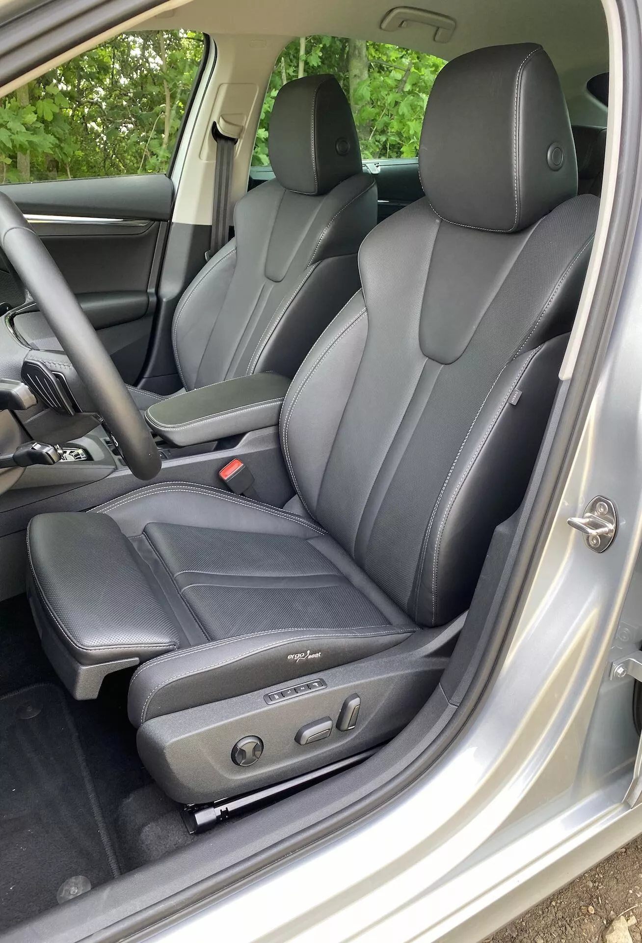 Interiérový pohľad na Škoda Octavia limuzínu 2020 zameraný na oblasť vodiča. Obsahuje tmavé kožené sedadlá s ergonomickým dizajnom a elektrickými ovládacími prvkami, viditeľné od ľavej strany cez otvorené dvere vodiča. Viditeľné opierky hlavy pre vodiča a zadných pasažierov naznačujú, že vozidlo pojme najmenej štyroch cestujúcich. Strieborné alebo svetlosivé obloženie interiéru kontrastuje so sedadlami a ovládací panel dverí obsahuje ovládače elektrických okien a zrkadiel, zdôrazňujúc technologický komfort a luxus.