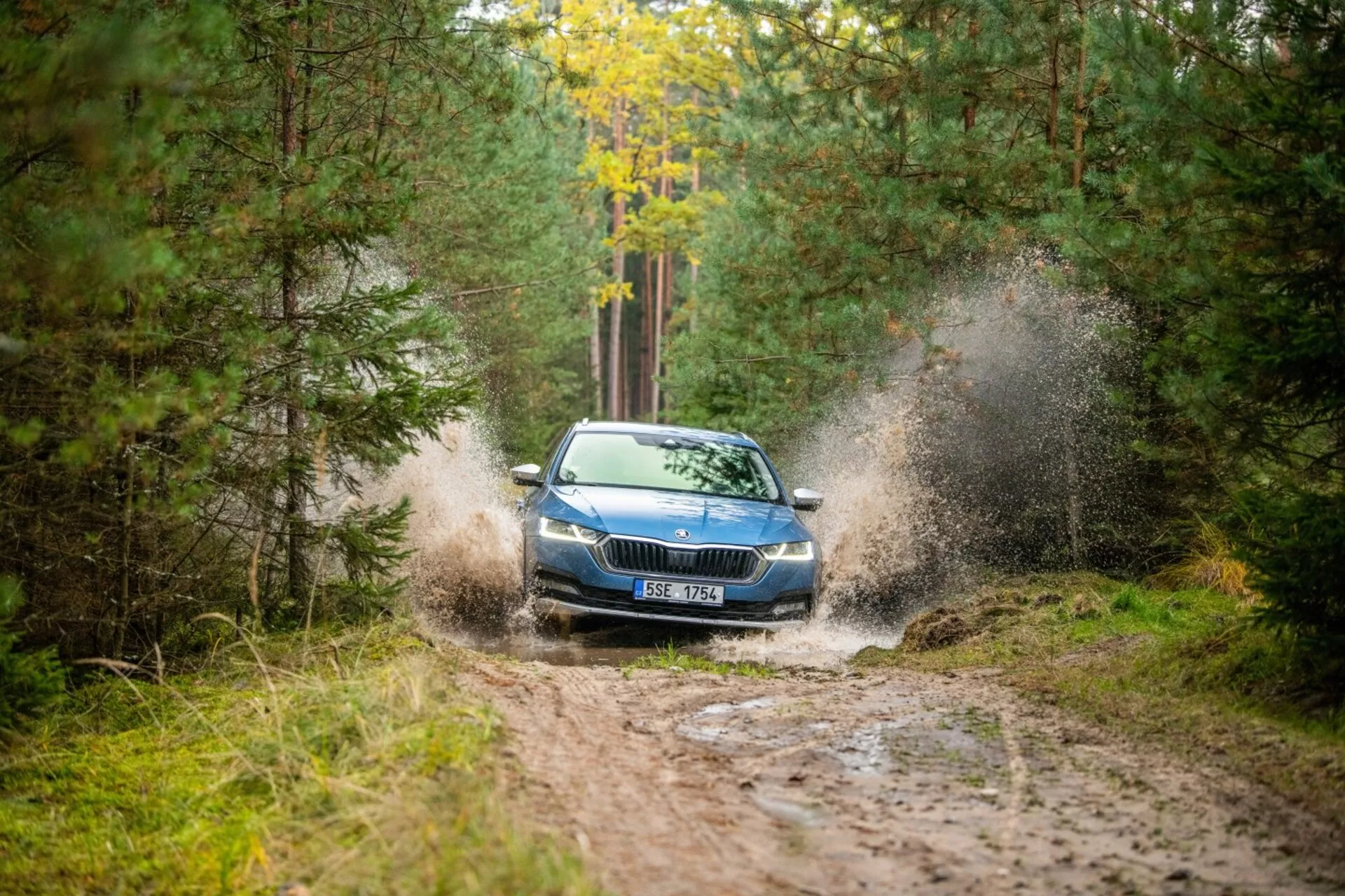 Limuzína Škoda Octavia z roku 2017 prechádza cez blátovú mláku v lesovej oblasti, čo zvýrazňuje jej dynamické vytváranie striekajúcej vody, prednú mrežu, svetlomety a kapotu s čiastočným výhľadom na strechu z uhla vpravo vpredu.