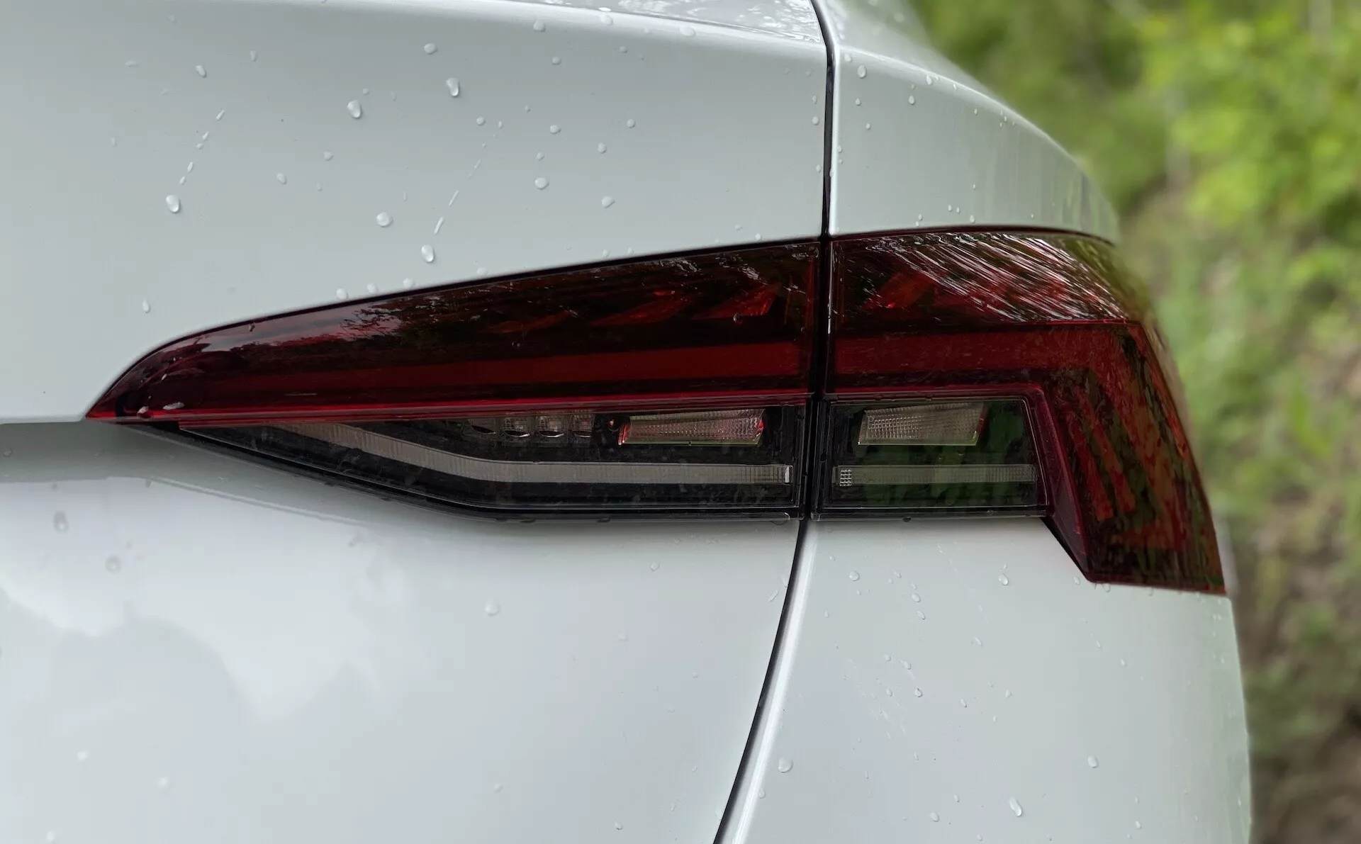 Zblízka zadná ľavá časť vozidla Škoda Octavia 2020 so zameraním na zadné svetlo. Obrázok je odfotený z diagonálneho uhla, ktorý zdôrazňuje moderný dizajn zadného LED svetla s ostrými uhlami a lesklým povrchom. Viditeľná je časť zadného kvadrantového panela a veka kufra, čo ukazuje plynulú integráciu a aerodynamickú estetiku. Hladký povrch s kvapkami vody naznačuje nedávne zrážky alebo čerstvé umytie.