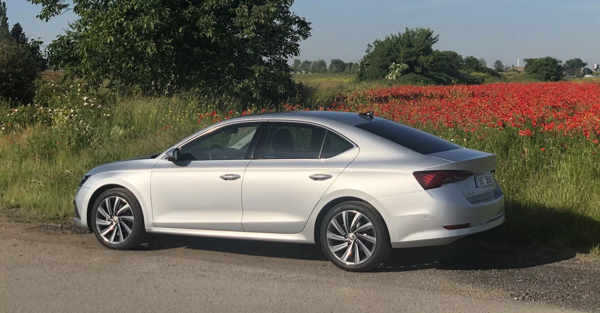 Bočný profil sedanu Škoda Octavia 2020 v striebornej farbe, zaparkovaného na štrkovej ceste s poľom a stromami v pozadí. Vozidlo má elegantnú, mierne klesajúcu strechu, ktorá prechádza do krátkej zadnej časti, typickej pre moderný sedan. Má štyri dvere, bočné zrkadlá v farbe karosérie a chrómové lišty okolo okien. Kolesá sú zliatinové s viacpaprskovým dizajnom a kľučky dverí sú tiež vo farbe karosérie. Pozdĺž boku je viditeľný jemný záhyb, ktorý prispieva k dynamickému vzhľadu. Senzory na bočných zrkadlách môžu naznačovať technológiu monitorovania slepého uhlu. Prostredie obsahuje výraznú škvrnu červených kvetov, pravdepodobne makov.