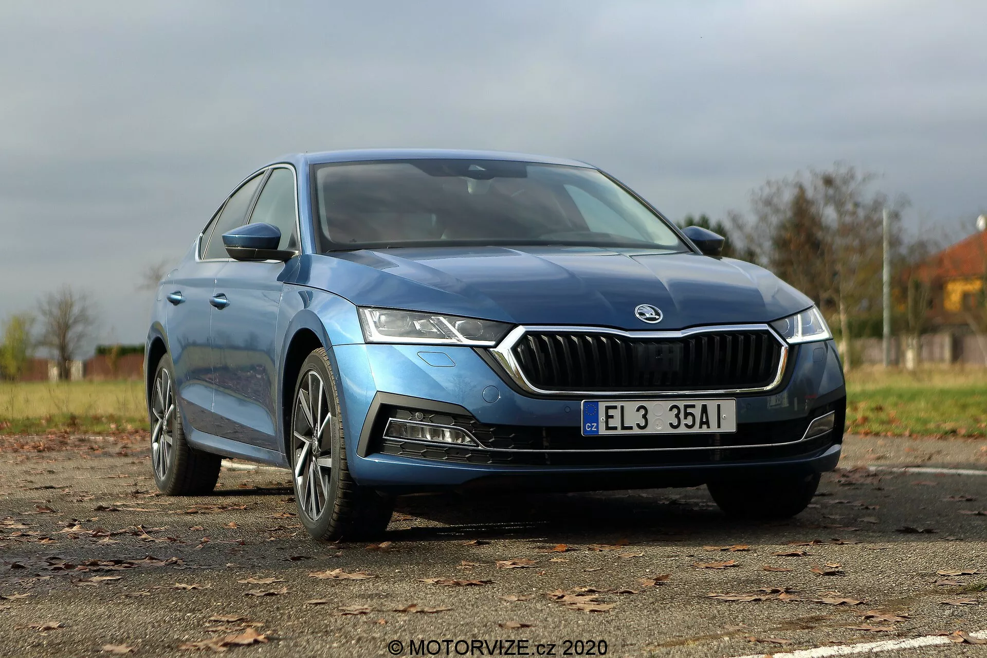 Obrázok modrej Škody Octavia Limousine 2020 zachytený z nízkeho trojštvrťového uhlu spredu, zdôrazňujúci výraznú chrómom orámovanú prednú mriežku, ostré LED svetlomety, kontúrovanú kapotu, moderné hliníkové kolesá a štýlové bočné zrkadlá s integrovanými smerovkami. Hladká karoséria a mierne tónované bezrámové okná naznačujú vyššiu úroveň výbavy, zatiaľ čo je prezentovaná celá dĺžka vozidla, vrátane viditeľných odznakov označujúcich model alebo typ motora.