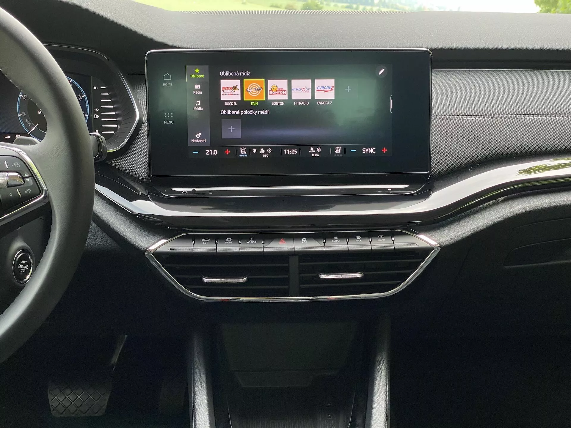 Interiér vozidla Škoda Octavia 2020 limuzína, s dôrazom na centrálnu konzolu a infotainment systém z perspektívy predného sedadla. Veľká, horizontálna dotyková obrazovka zobrazuje farebné používateľské rozhranie s ikonami pre navigáciu, prehrávanie médií a nastavenia vozidla. Pod dotykovou obrazovkou sú elegantné ventilačné otvory s prevažne chrómovým lemovaním, nasledované fyzickými tlačidlami a otočnými regulátormi pre klimatizáciu. Palubná doska a konzola obsahujú kombináciu textúr podobných koži a matných a lesklých plastov, čo naznačuje vysokokvalitný dizajn pre stredný až vyšší trhový segment.