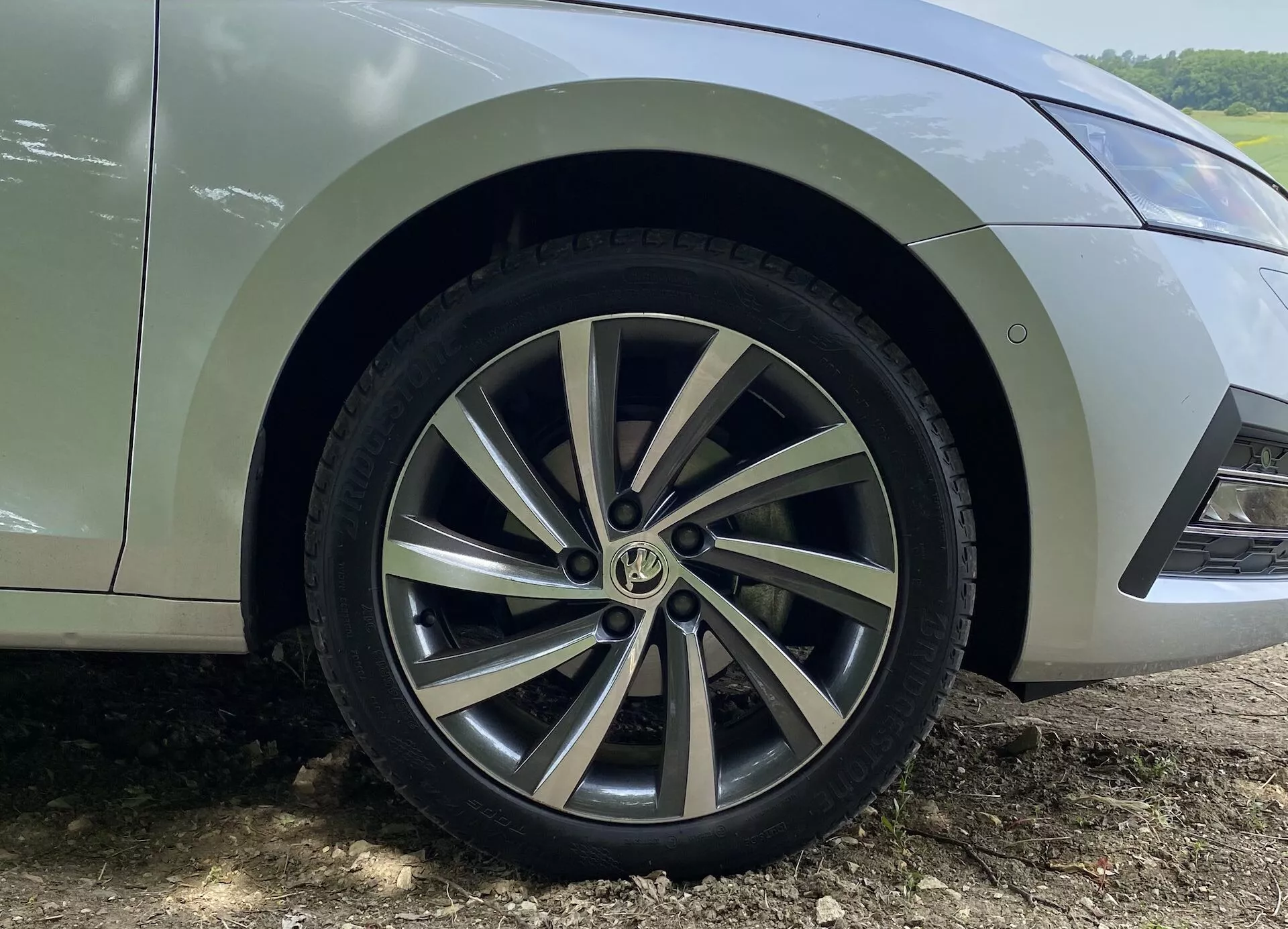 Detailný záber na predné koleso a spodný blatník vozidla Škoda Octavia Limousine 2020, zvýrazňujúci dizajn viaclúčového hliníkového kolesa s kombináciou striebornej farby a opracovaných plôch. Viditeľný dezén pneumatiky ukazuje malé opotrebovanie. Auto je zaparkované na hlinenom povrchu s blatom za kolesom, čo naznačuje nedávnu jazdu v teréne alebo na nezpevnených cestách. Zobrazená je iba časť auta od spodných predných dverí po predný nárazník, zachytávajúci detaily kolesa a blatníka z nízkeho uhla.