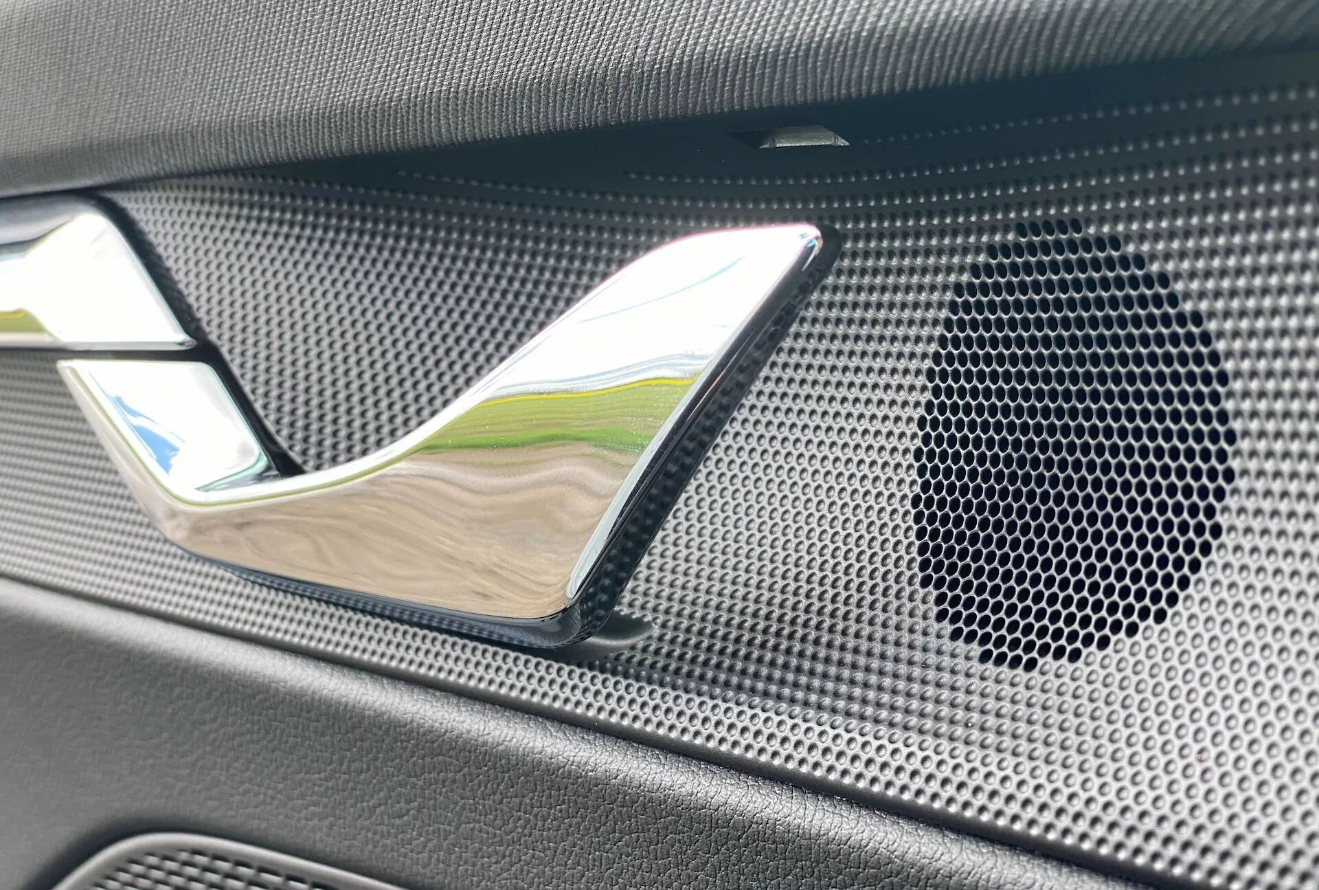 Detailný záber interiéru modelu 2020 Skoda Octavia limuzína so zvýraznením chrómovanej kľučky dverí a reproduktorovej mriežky s voštinovým vzorom a textúrovaným pravdepodobne karbónovým materiálom, zdôrazňujúci luxus a vysokokvalitný audiosystém.