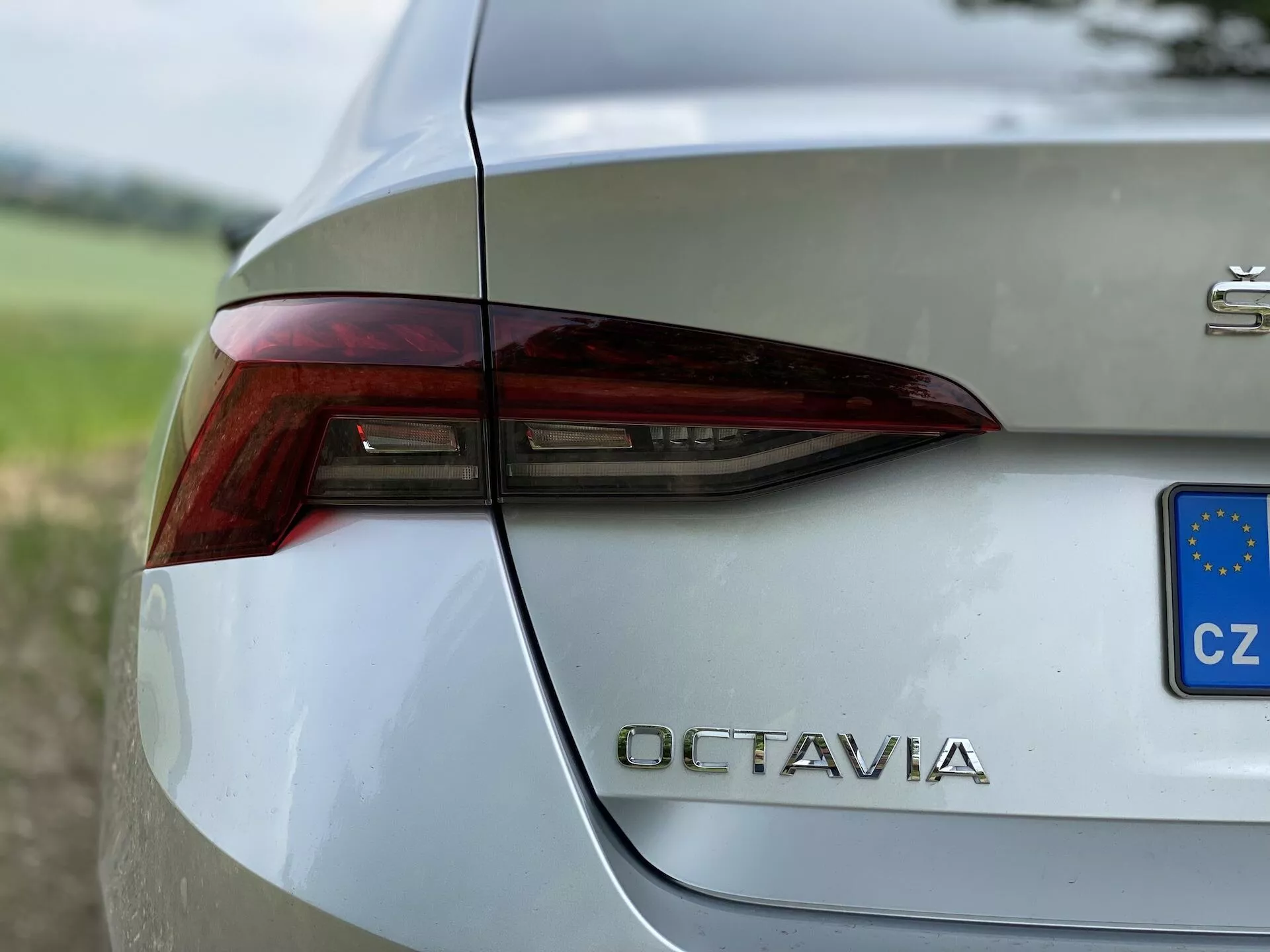 Detailný pohľad na zadnú pravú časť strieborného Škoda Octavia 2020. Obrázok zachytáva ostrý a hranatý dizajn zadného svetla s integrovanými prvkami brzdových svetiel, smeroviek a pravdepodobne cúvacích svetiel. Zadné veko kufra zreteľne zobrazuje označenie modelu 'OCTAVIA' chrómovými písmenami pod zadným svetlom. Malý modro-žltý obdĺžnik s hviezdičkami naznačuje registráciu Česká republika. Leštený, kovovo-čerpaný povrch karosérie odráža okolie, ktoré je umiestnené na rozmazanom pozadí zelene, čo zvýrazňuje sofistikovaný dizajn vozidla.