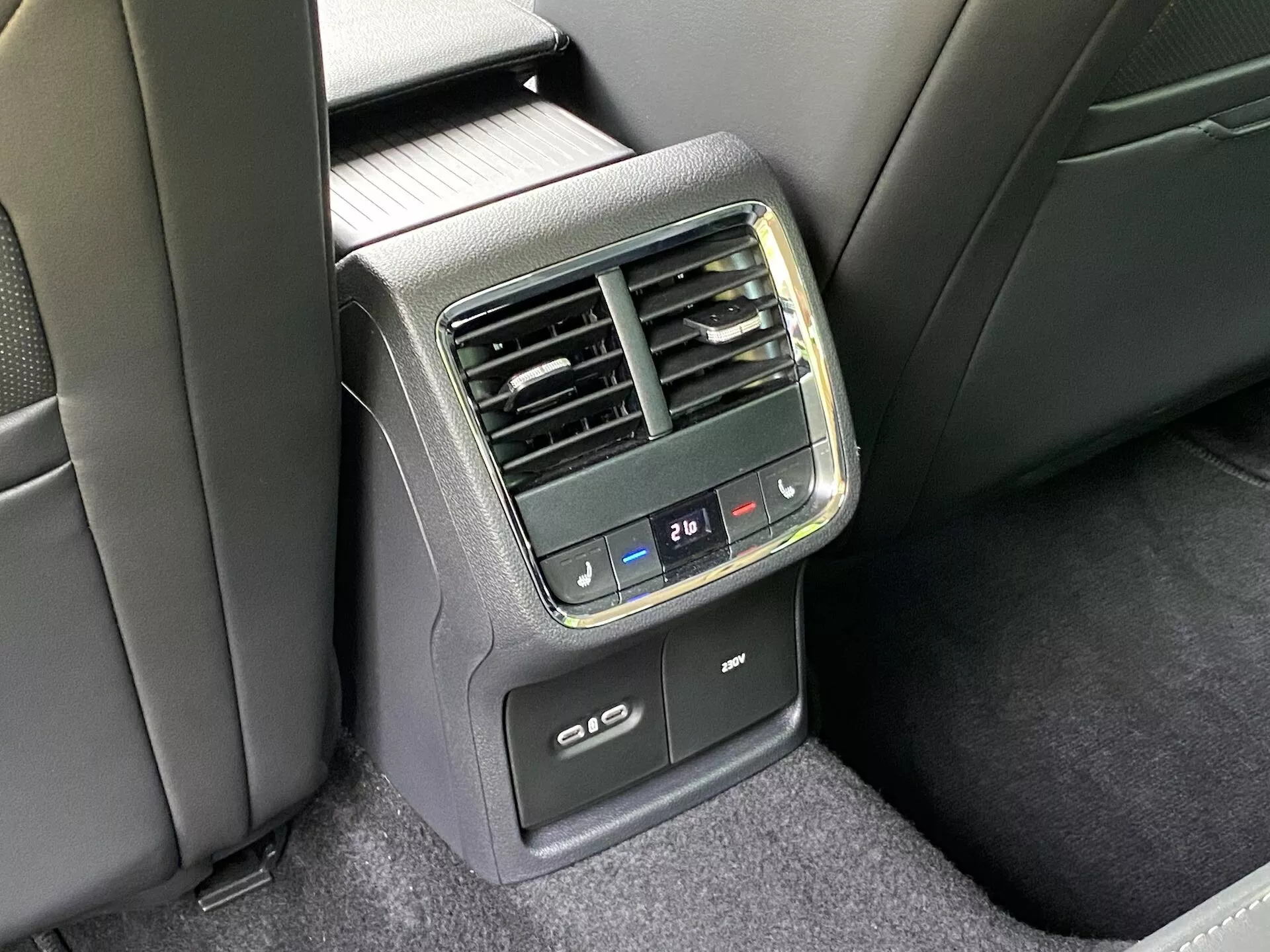Detailný pohľad na zadný ovládací panel klimatizácie v zadnej časti automobilu Skoda Octavia 2020. Záber z offsetového uhla ponúka jasný pohľad na stredový panel na zadnej strane prednej lakťovej opierky. Obsahuje tlačidlá digitálneho ovládania klimatizácie s nastavením teploty, vertikálne ventilačné otvory, digitálny displej teploty nastavený na 21℃, USB porty na nabíjanie a potenciálnu funkciu vyhrievania alebo chladenia zadných sedadiel. Materiály sú kombináciou plastu a látky v čiernej a tmavosivej farebnej schéme pri prirodzenom osvetlení.