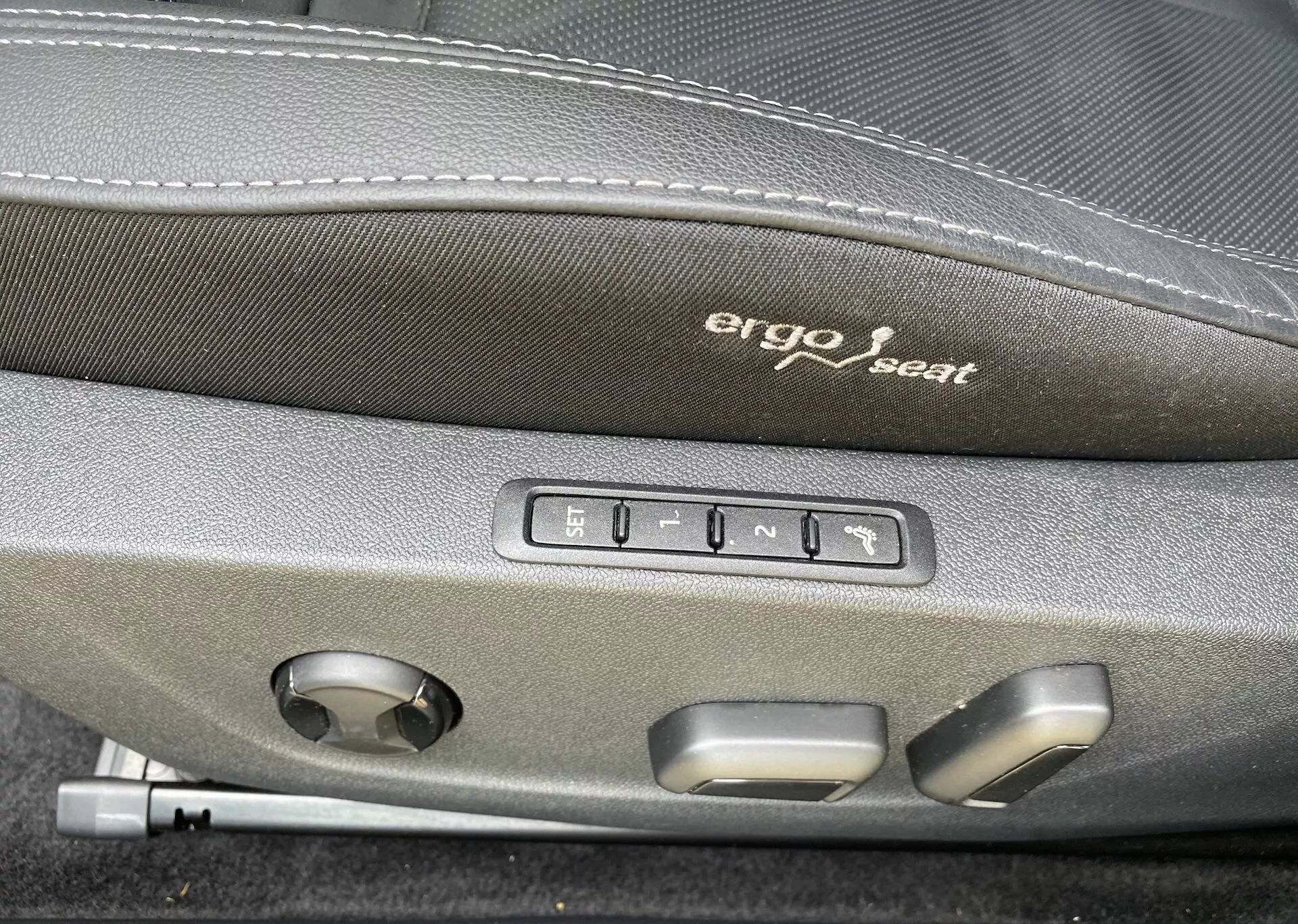 Detailný záber interiéru vozidla Škoda Octavia 2020, zameraný na ergonomické ovládanie sedadla. Na tmavšej hornej časti sedadla je vyšité slovo 'Ergo seat'. Pod týmito slovami je vidieť päť tlačidiel: jedno pre pamäťovú funkciu sedadla a štyri pre nastavenie polohy sedadla dopredu, dozadu, hore, dole a náklon operadla. Tiež je tu okrúhly volič, pravdepodobne pre nastavenie bedrovej opierky alebo iné multifunkčné úpravy sedadla, čo naznačuje vyššiu úroveň komfortu a pokročilú technológiu v tomto vozidle.