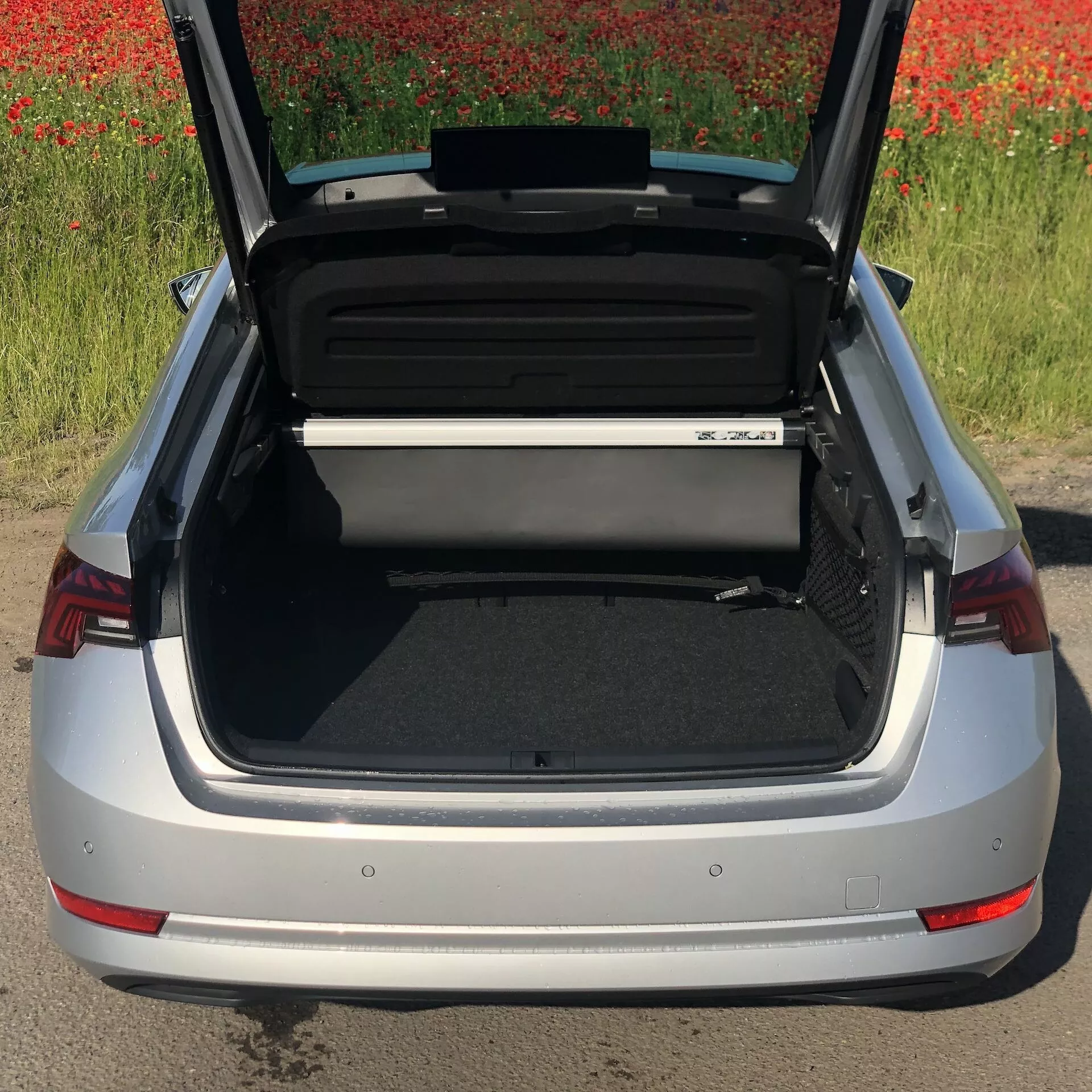 Pohľad z vysokého uhla na strieborný sedan Skoda Octavia 2020 s otvoreným kufrom, ktorý ukazuje priestranný a organizovaný úložný priestor. Viditeľný je zadný nárazník, charakteristické červené zadné svetelné klustery a časť emblému automobilu, s poľom červených kvetov v pozadí.