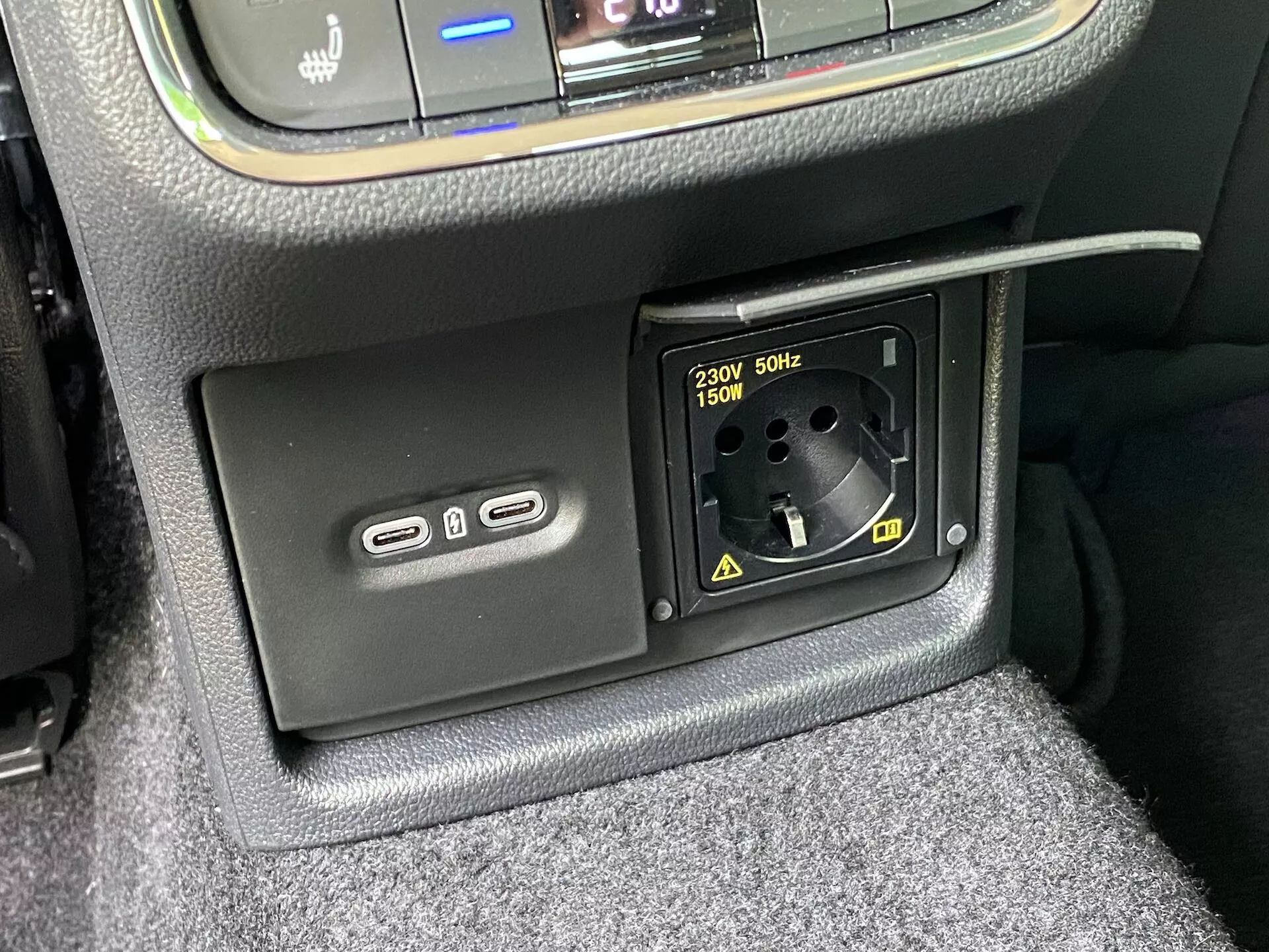 Detailný pohľad na funkcie napájania v interiéri vozidla Škoda Octavia 2020: spodná časť stredovej konzoly s dvoma portami USB-A na ľavej strane na nabíjanie alebo pripojenie k multimediálnemu systému a zásuvkou na 230 V AC pre európske zástrčky s podporou až 150 W na pravej strane, obklopená textúrovaným obložením a viditeľným kobercom.