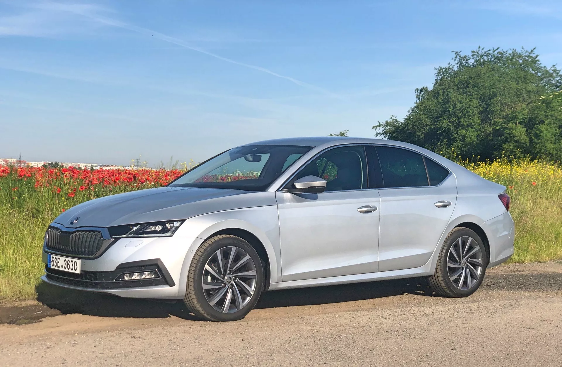 Obrázok zobrazuje štvordverový strieborný sedan, konkrétne Škoda Octavia 2020 (Octavia-Limousine), zaparkovaný vedľa cesty s prírodným pozadím tvoreným polí a radom stromov. Fotografovaný z bočného profilu trochu spredu, automobil má viacramenné zliatinové kolesá, elegantné LED svetlomety a výraznú mriežku s vertikálnymi lamelami. Moderný dizajn zahŕňa tvarované línie karosérie a klesajúcu strešnú líniu s bočnými zrkadlami integrovanými smerovými svetlami. Viditeľné sú tiež zadná anténa a senzory na prednom nárazníku, ktoré naznačujú pokročilé asistenčné systémy pre vodiča.