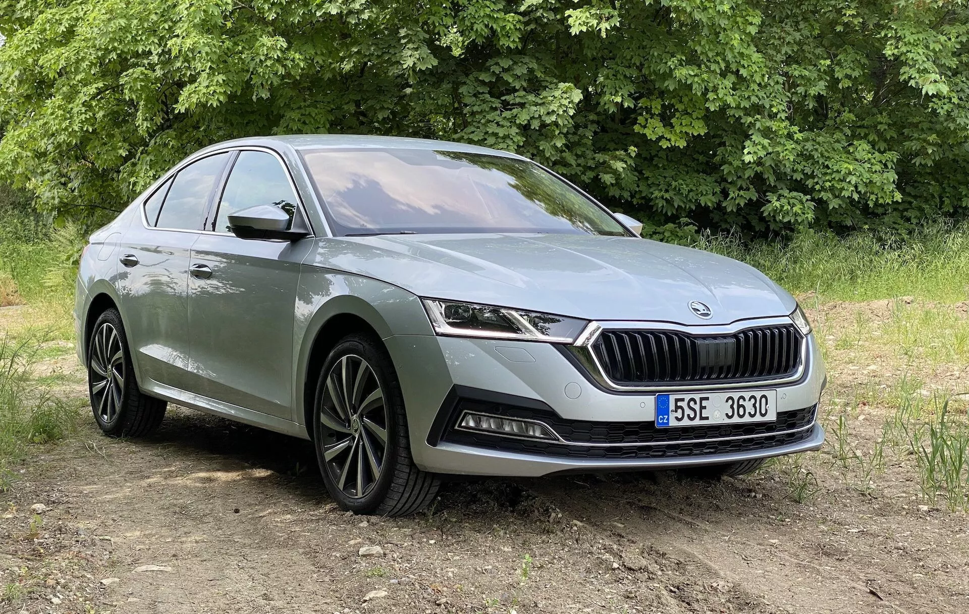 Trojštvrťový predný pohľad na moderný sedan Škoda Octavia 2020, zobrazený z mierne nižšieho uhla, zameraný na prednú fasádu, mriežku s vertikálnymi lamelami a výrazným logom, elegantné vodorovne orientované LED svetlomety a dolný nárazník s hmlovými svetlami a prívodmi vzduchu. Auto je umiestnené na nezpevnenom povrchu s vegetáciou v pozadí, za prirodzeného denného svetla. Ďalšie viditeľné detaily zahŕňajú ráfiky z ľahkých zliatin s viaclamelovým dizajnom, charakteristickú líniu bežiacu pozdĺž profilu vozidla a kľučky dverí. Vhodné na predstavenie automobilového dizajnu a moderných automobilových technológií v vonkajšom prostredí.