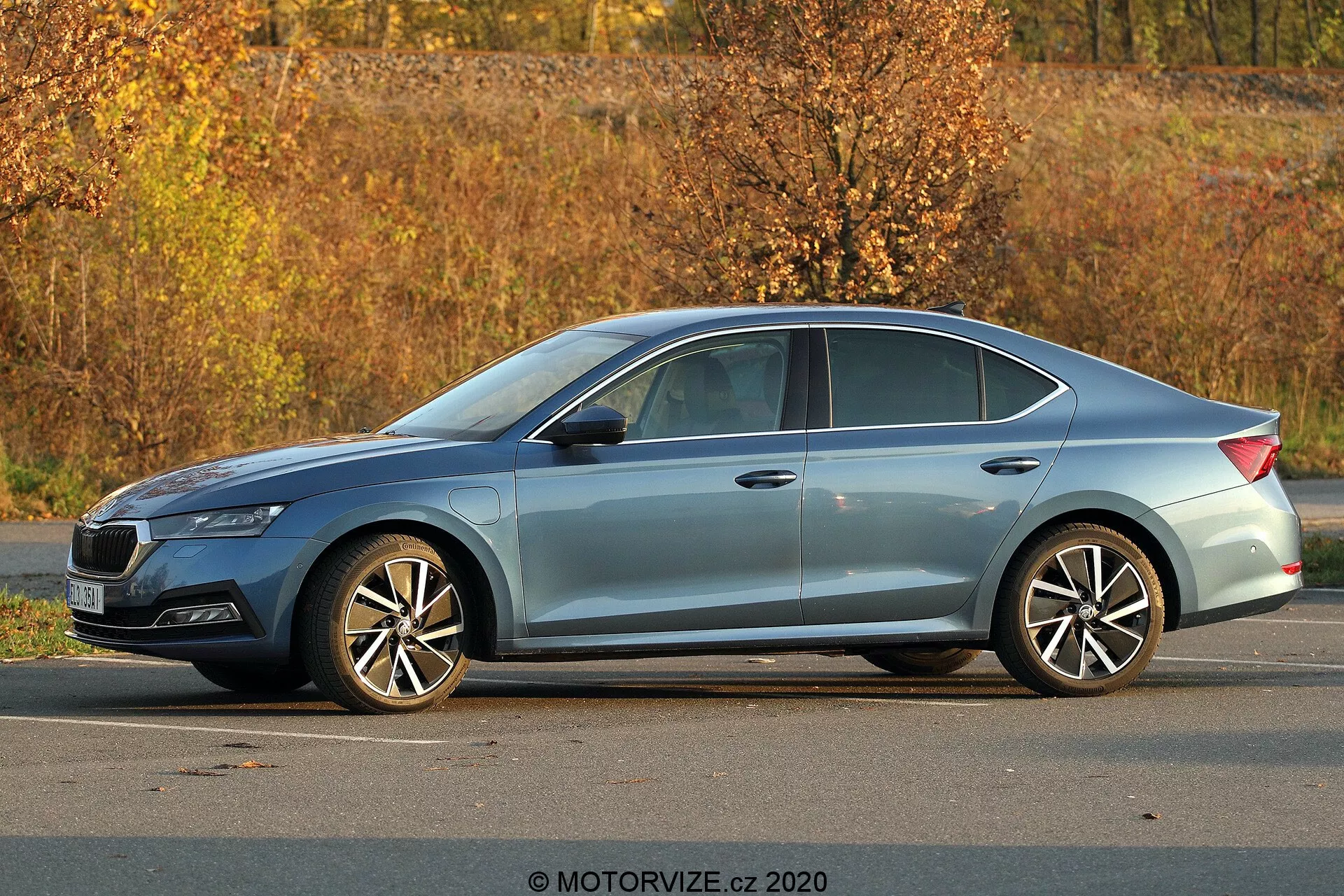 Moderný sedan Skoda Octavia 2020 zaparkovaný vonku, pohľad zboku z troch štvrtín ukazujúci plný profil, dĺžku a výšku; viditeľné detaily zahŕňajú ostré svetlomety, klesajúcu líniu strechy, charakterové línie, okná s chrómovými rámami, viacramenné zliatinové kolesá, jesenné prostredie s listami a prirodzené denné osvetlenie.