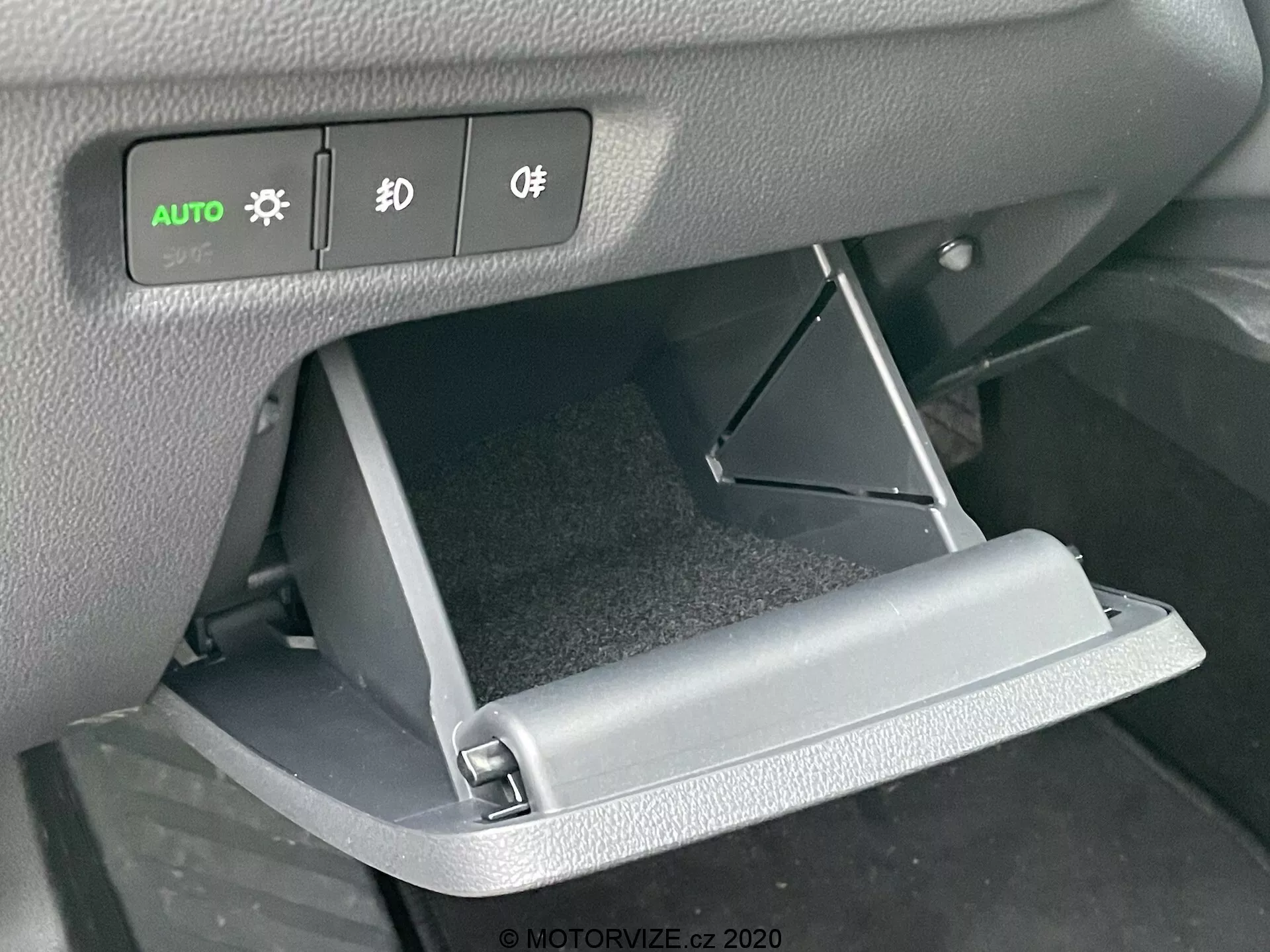 Detailný záber interiéru vozidla Škoda Octavia 2020 zameraný na otvorenú priehradku na rukavice a ovládacie prvky na palubnej doske. Predný uhol zdôrazňuje sklopené dvierka priehradky a osvetlené tlačidlá svetlometov, hmly a ovládania osvetlenia nad ňou.