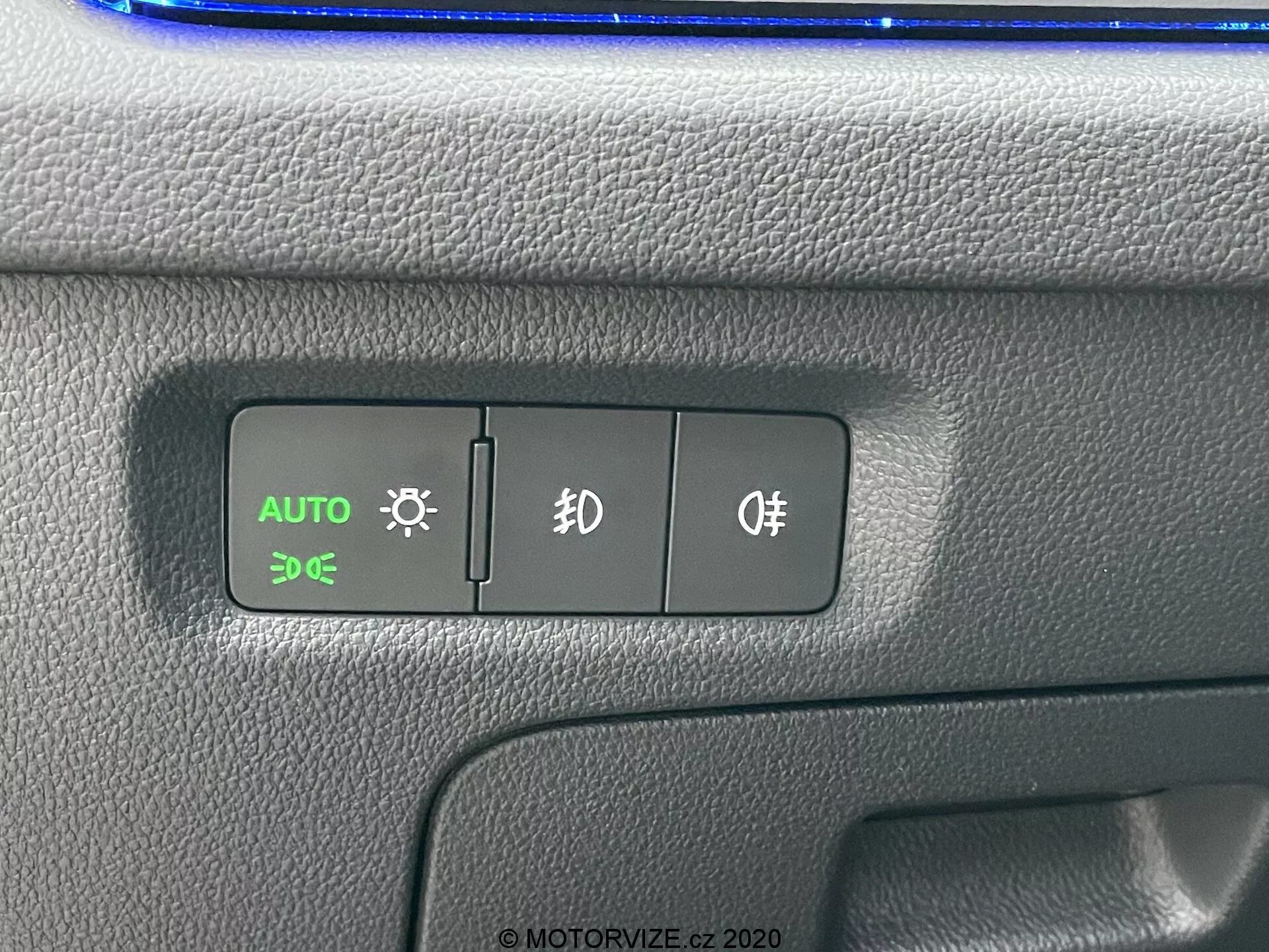 Detailný záber na ovládací panel vozu Škoda Octavia 2020, so zameraním na spínače automatických svetiel, parkovacích svetiel a hmloviek. Tlačidlo AUTO je osvetlené na zeleno, čo naznačuje aktívne automatické ovládanie svetiel. Textúrovaný plastový panel má dvojfarebný dizajn s možným modrým odrazem na vrchu, v nastavení interiéru vozidla.