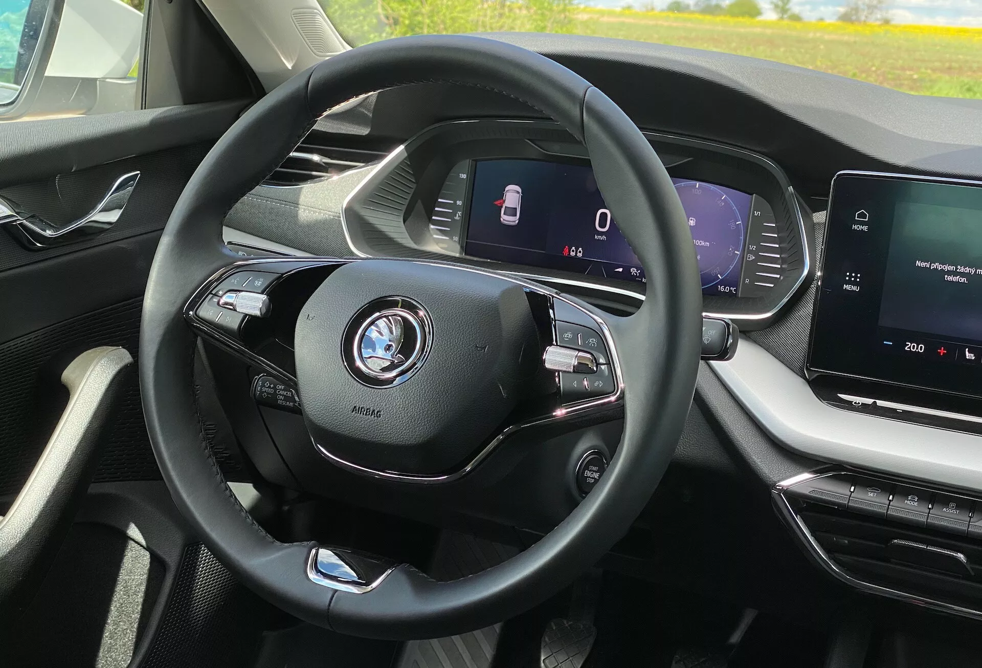 Pohľad do interiéru vozidla Škoda Octavia 2020 z perspektívy sedadla vodiča; moderný trojramenný volant so multifunkčnými ovládačmi pre médiá a telefón, viditeľné logo automobilky; plne digitálny prístrojový panel, ktorý zobrazuje rýchlosť a bezpečnostné funkcie ako asistent udržania v jazdnom pruhu; stredová konzola s dotykovou obrazovkou infotainmentu, zobrazuje mediálny systém a možnú navigáciu; moderný, luxusný interiér s čistými líniami a dobre navrhnutými vetracími otvorami; žiadny výhľad na exteriér, len bezprostredné okolie vodiča v kabíne; náznaky pokročilých technológií automobilu ako adaptívny tempomat a funkcie konektivity.