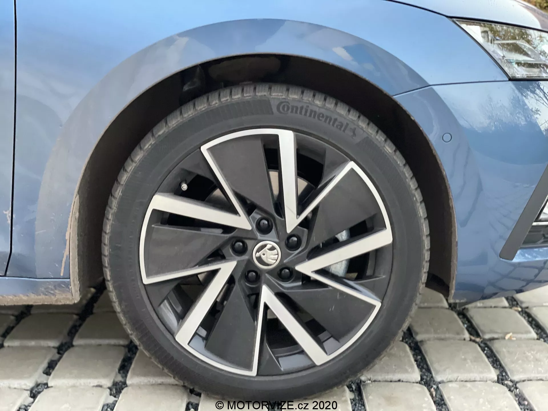 Detailný záber na predné koleso a čiastočný pohľad na predný blatník modelu Škoda Octavia 2020 limuzíny. Koleso má moderný, viaclúčový dizajn z ľahkých zliatin v dvojfarebnej čiernej a striebornej farbe s logom výrobcu uprostred. Pneumatika je značky Continental, pravdepodobne z rady Conti, s čiastočne viditeľnými alfanumerickými špecifikáciami veľkosti. Za lúčmi je vidieť brzdový kotúč a možno aj brzdové strmene, čo naznačuje moderný brzdový systém. Karoséria je znázornená v metalickej modrej farbe.