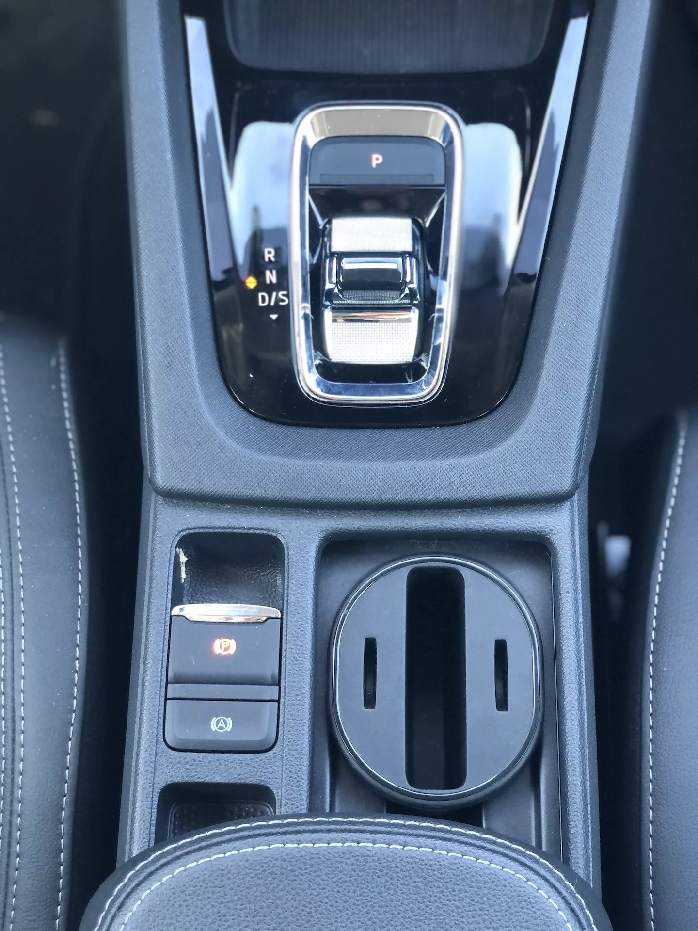 Detailný pohľad na oblasť automatickej prevodovky v modeli Škoda Octavia Limousine 2020. Radiaca páka je strieborná a čierna elektronická páka s možnosťami P, R, N a D/S. Vľavo sú dve ovládacie tlačidlá, pričom horné je osvetlené červenou farbou, pravdepodobne ide o elektronickú parkovaciu brzdu. Vpravo je zakrytá úložná priehradka s dvoma držiakmi na poháre. Čierna konzola má koženkovú textúru s detailným prešívaním, čo naznačuje luxusnejší interiér s pokročilým prevodovým systémom a elektronickými parkovacími systémami.