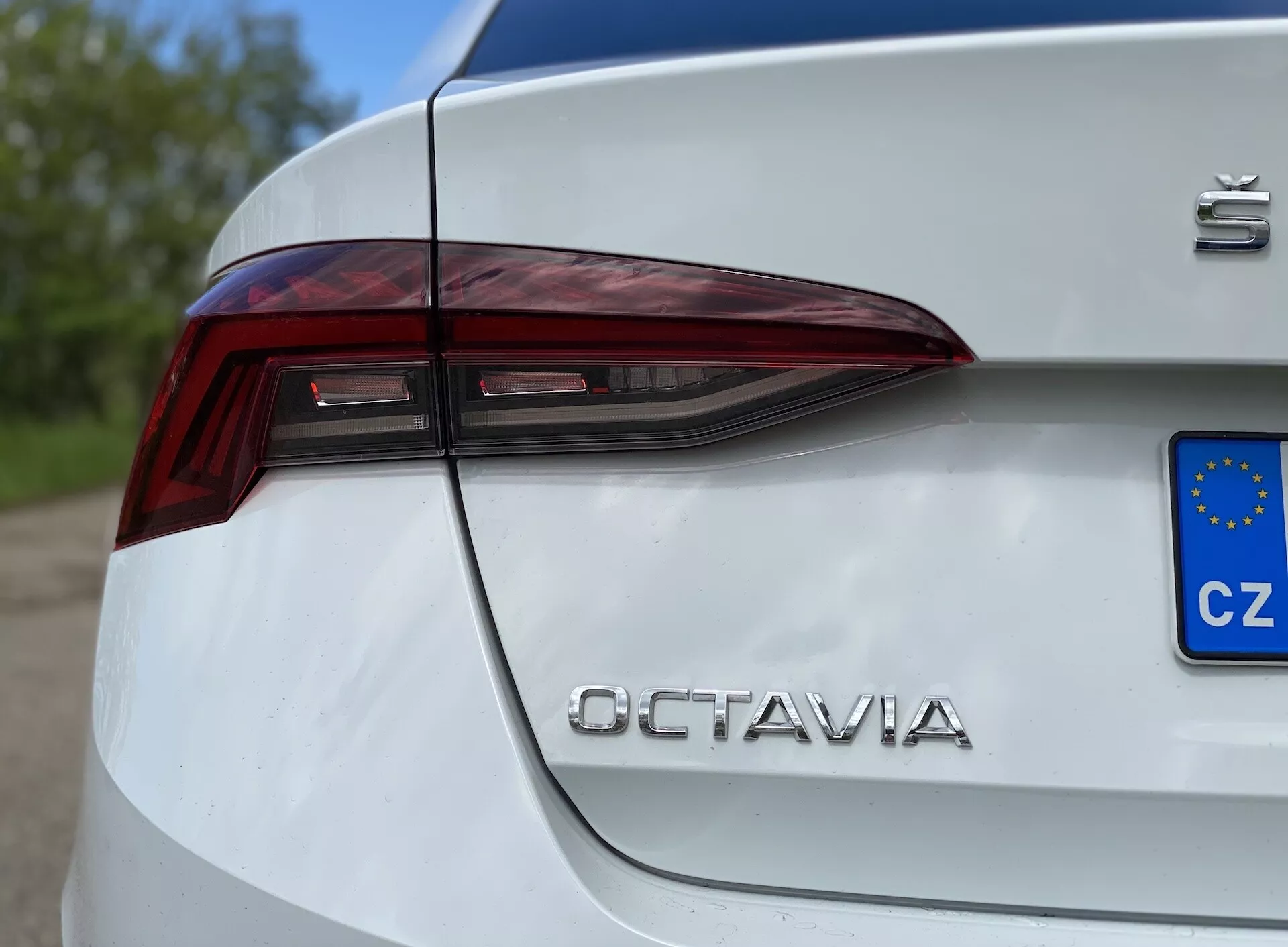 Detailný bočný pohľad na zadnú štvrtku bielej Škody Octavia Limousine 2020, zdôrazňujúci ostrý, uhlový dizajn LED zadných svetiel, ktoré prechádzajú na kufor, spolu so znakom 'Škoda' a chrómovým nápisom 'OCTAVIA', čiastočne viditeľnou európskou poznávacou značkou (CZ), zaznamenaný z nízkeho uhla.