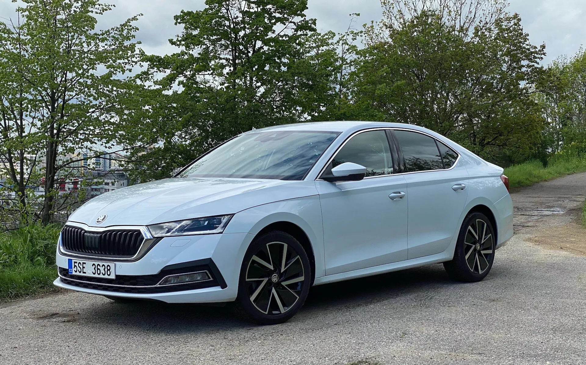 Mierne šikmý bočný pohľad na svetlomodrý sedan Škoda Octavia 2020, umiestnený pri ceste s pozadím zeleného lístia. Ukazuje moderný dizajn s elegantnou štvordverovou karosériou, výraznou mriežkou, agresívnymi LED svetlometmi, veľkými viaclúčovými zliatinovými kolesami, bočnými zrkadlami vo farbe karosérie s integrovanými smerovkami, klesajúcou strechou smerom dozadu, tónovanými oknami s chrómovou lištou, zapustenými kľučkami dverí, nízkoprofilovými pneumatikami a odznakom označujúcim model alebo technologický balíček.