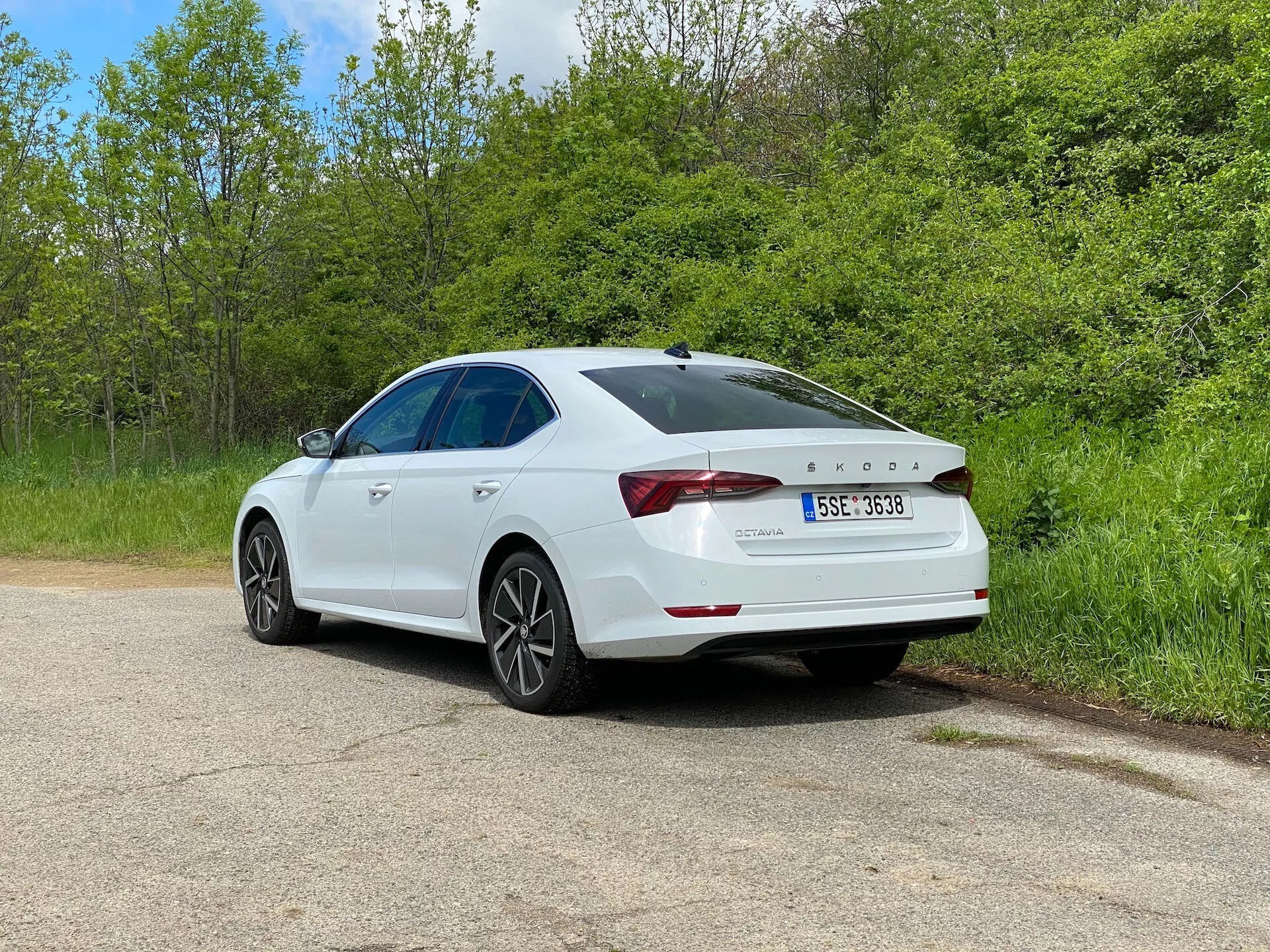 Biely sedan Skoda Octavia 2020 v trojštvrtečnom zadnom pohľade, zaparkovaný na štrkovej ceste, obklopený zeleňou, s detailným pohľadom na charakteristické zadné svetlá, zadný spojler, zliatinové kolesá a viditeľné strešné okno, zdôrazňujúci jeho moderný dizajn a technologické prvky vrátane parkovacieho senzora na nárazníku.